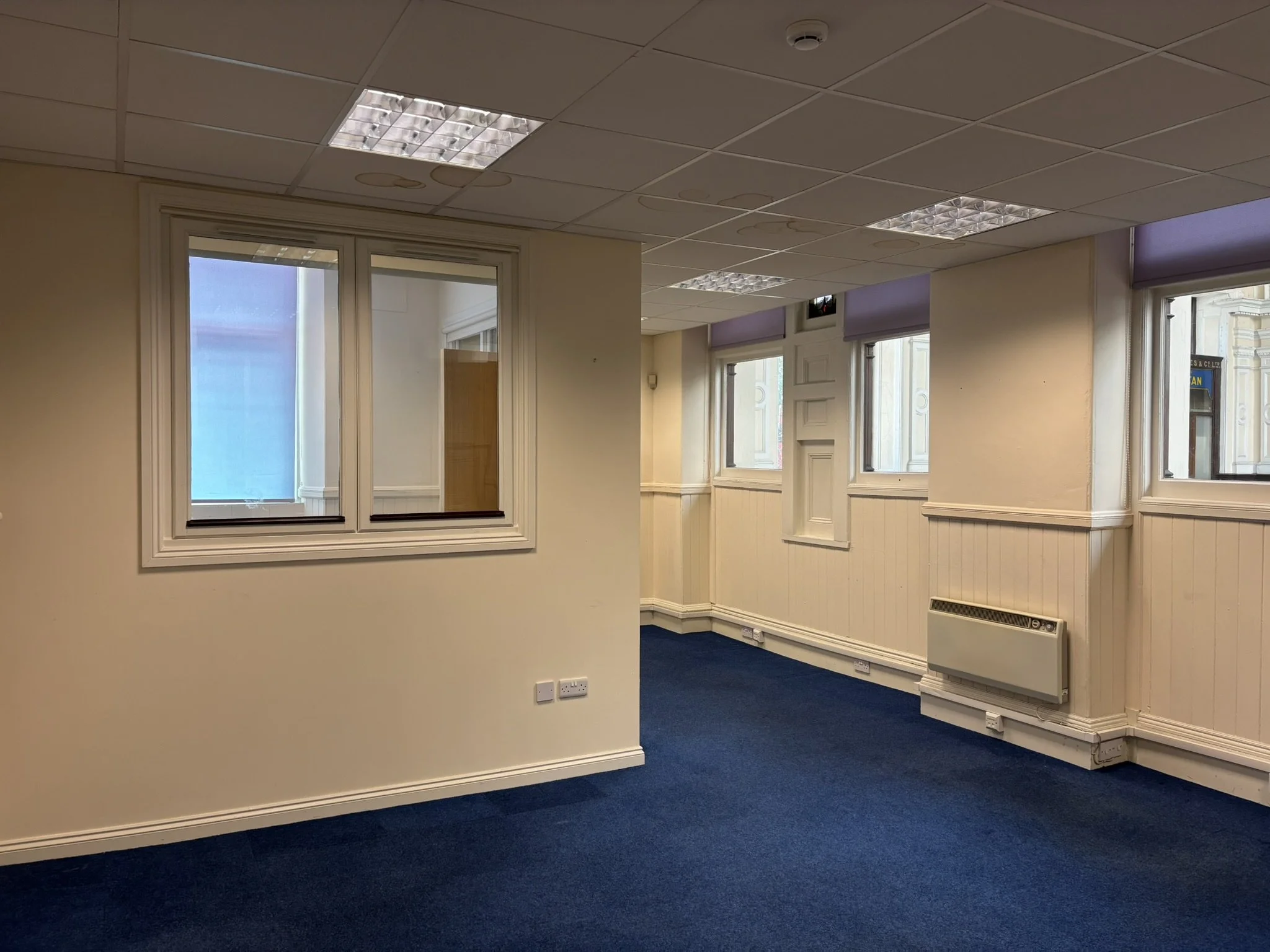 Empty office space with beige walls, multiple windows with purple blinds, ceiling lights, and blue carpet flooring.