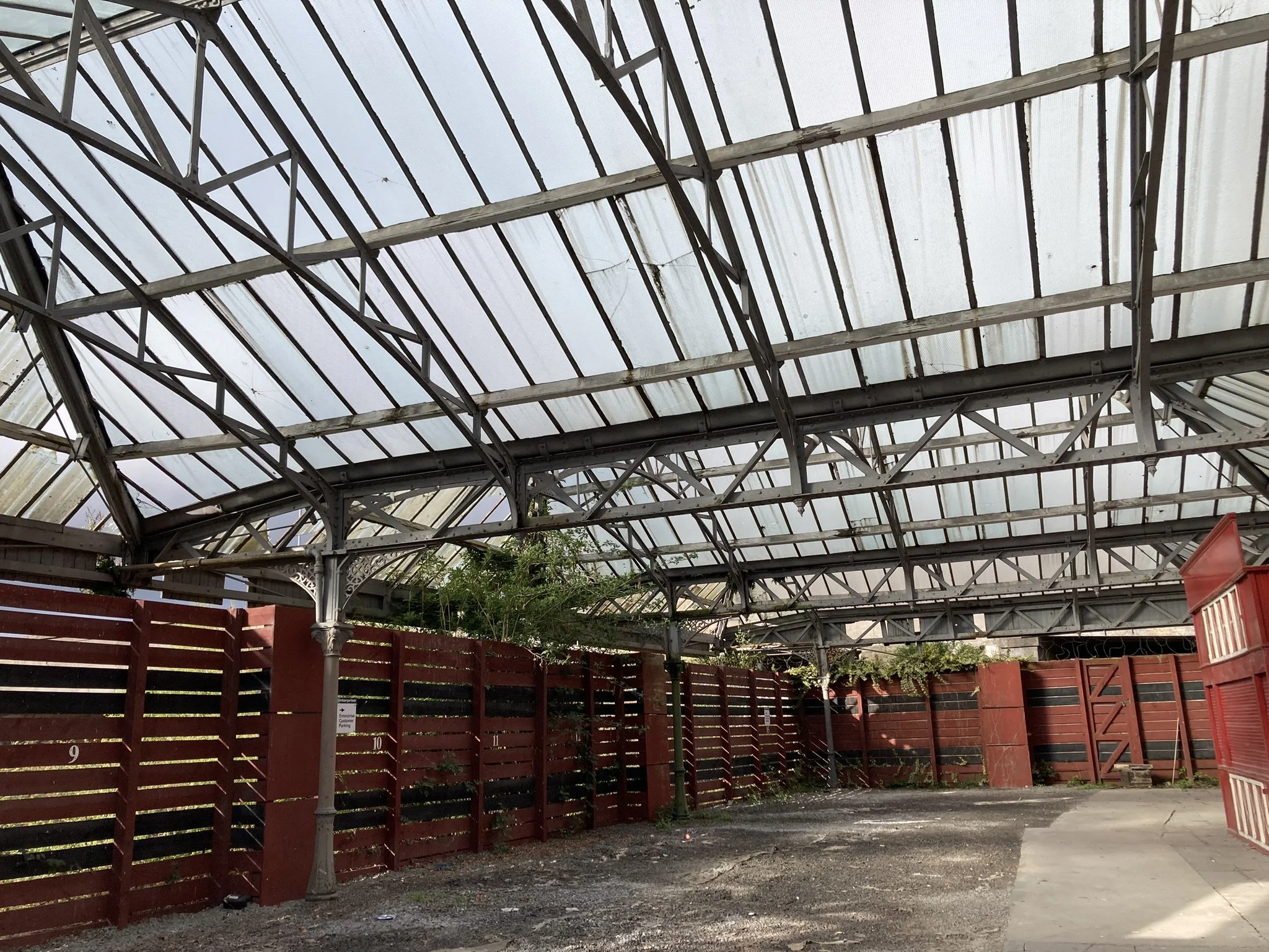 View of a covered outdoor area with a metal and glass roof structure with red wooden fencing around the perimeter and a few small trees.