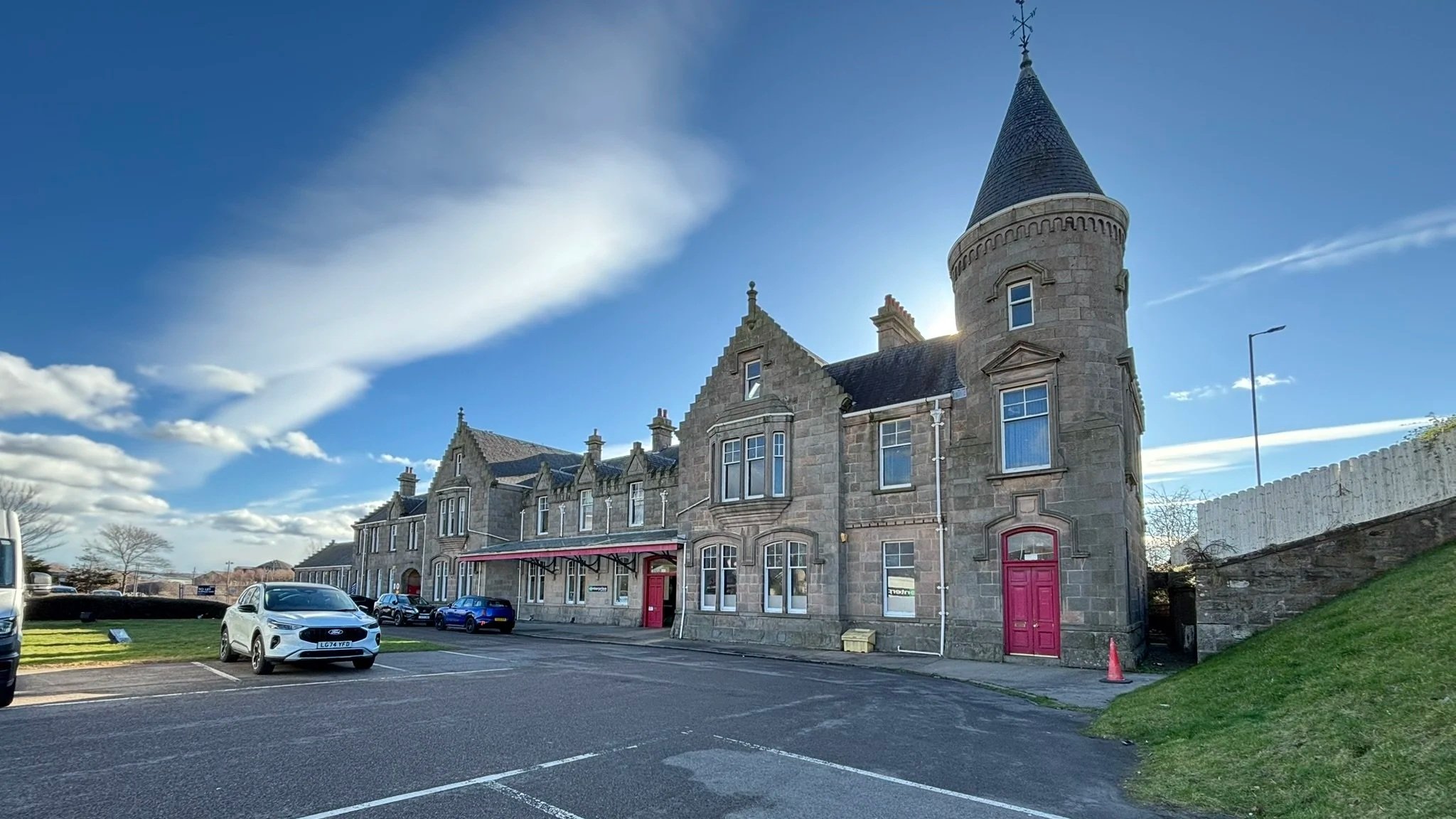 A historic stone building with a round tower and a pointed roof, adjacent to a parking lot with cars, under a partly cloudy sky during daytime.