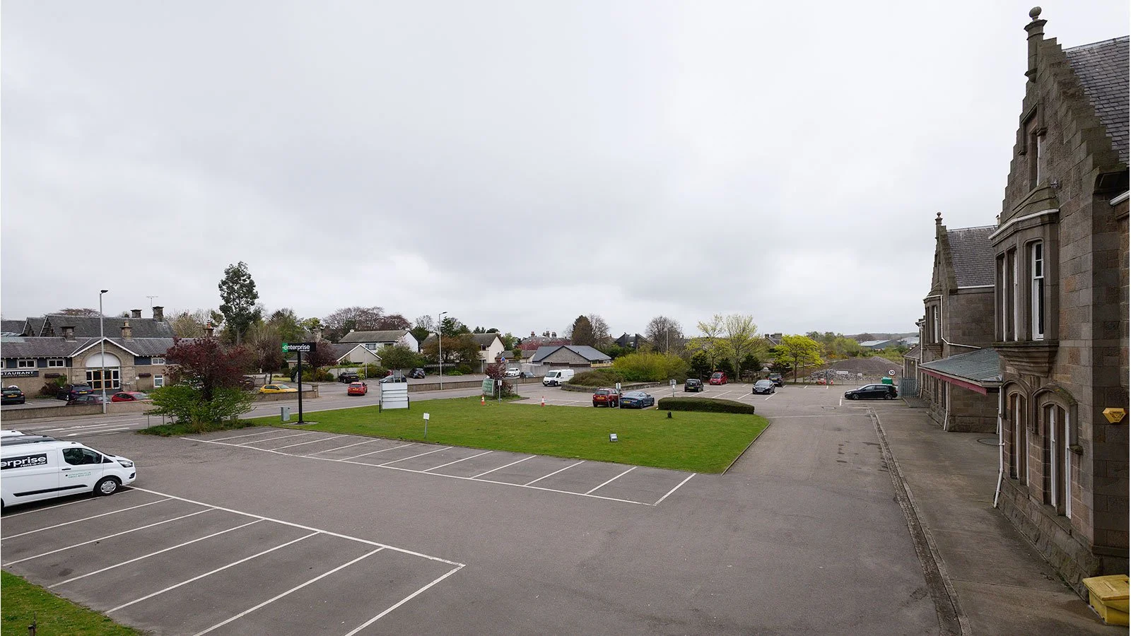A parking lot with several empty spaces, bordered by a grassy area with trees, and a street with cars and houses in the background under a cloudy sky.