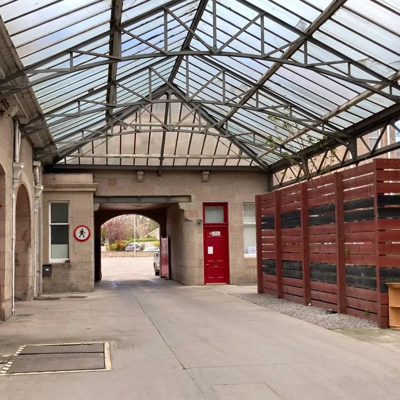 A covered walkway in a parking lot area with a stone building, a red door, and a wooden fence on the right side.