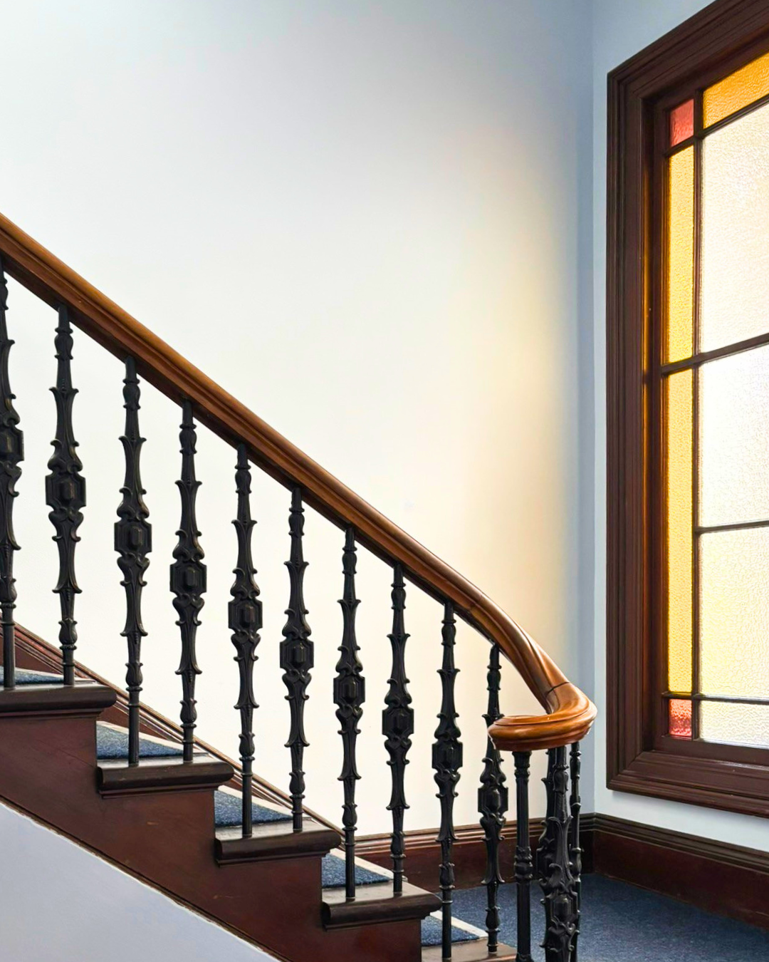 Interior view of a staircase with a wooden handrail, black decorative balusters, and a stained glass window with yellow, red, and orange panes.