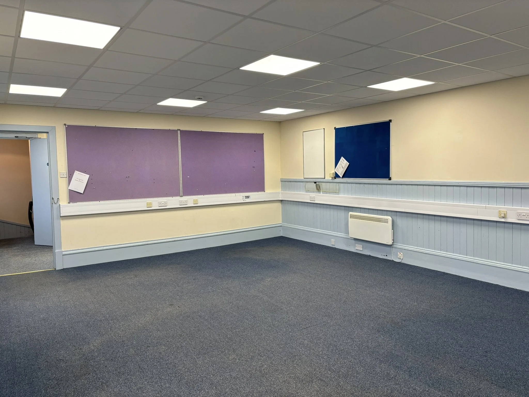 Empty classroom with beige walls, purple and blue bulletin boards, gray carpet, and fluorescent ceiling lights.