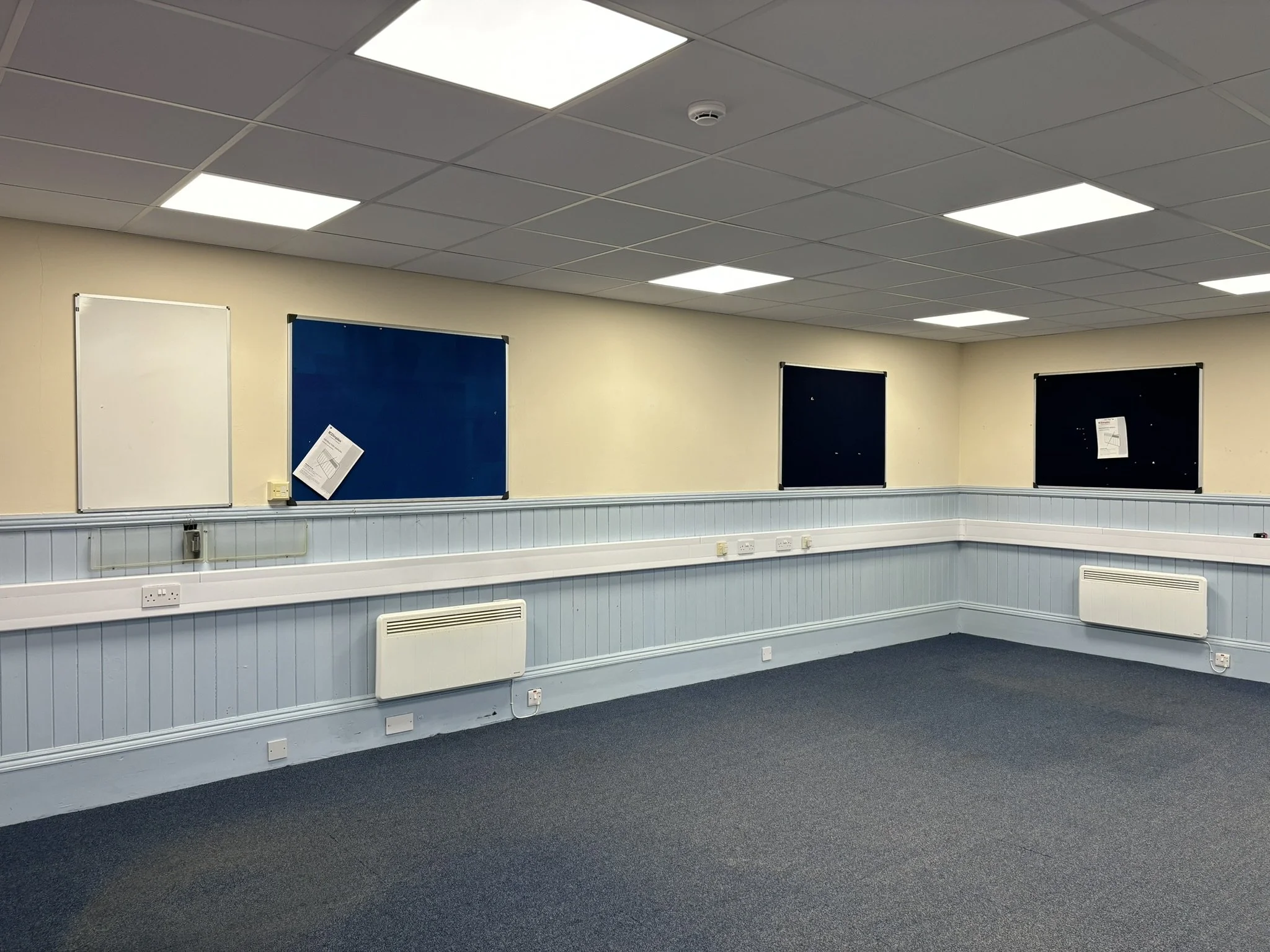 A room with beige walls, blue and white notice boards, and a dark blue carpet. The ceiling has square white LED lights and a smoke detector.