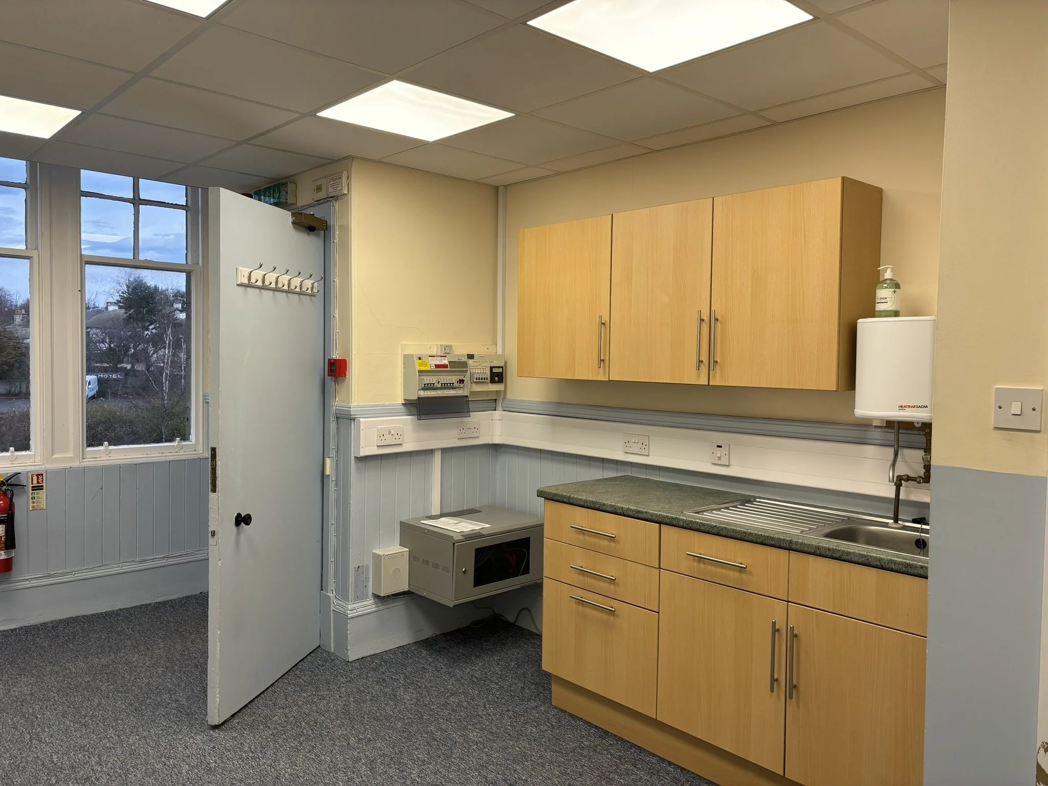 Interior of a room with beige walls and a ceiling with fluorescent lights. There are wooden cabinets, a stainless steel sink, and a countertop. A window shows a cloudy sky and trees outside. Fire safety equipment, electrical outlets, and a bottle of 