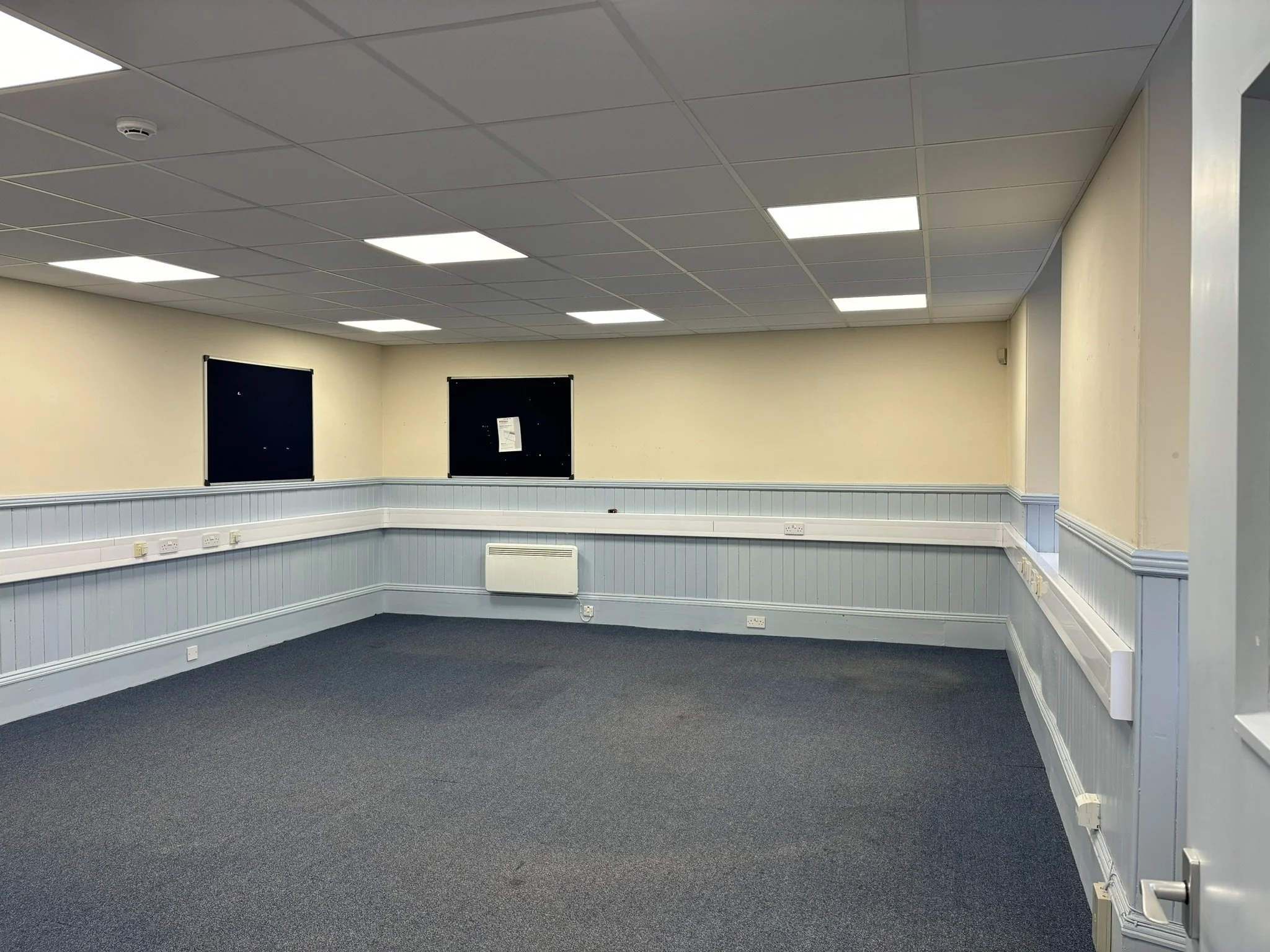 Empty room with beige walls, a blue-carpeted floor, white wainscot paneling, two black notice boards, and ceiling lights.