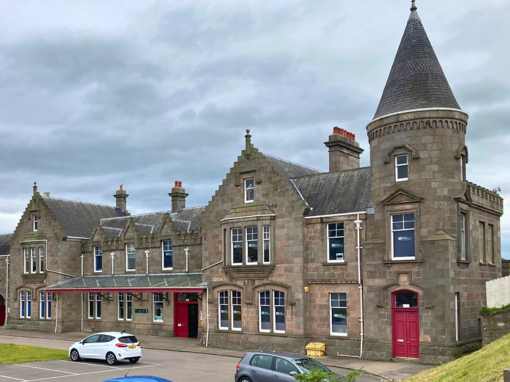 A historic stone building with turrets and red doors, situated in a parking lot with a few cars. Overcast sky above.
