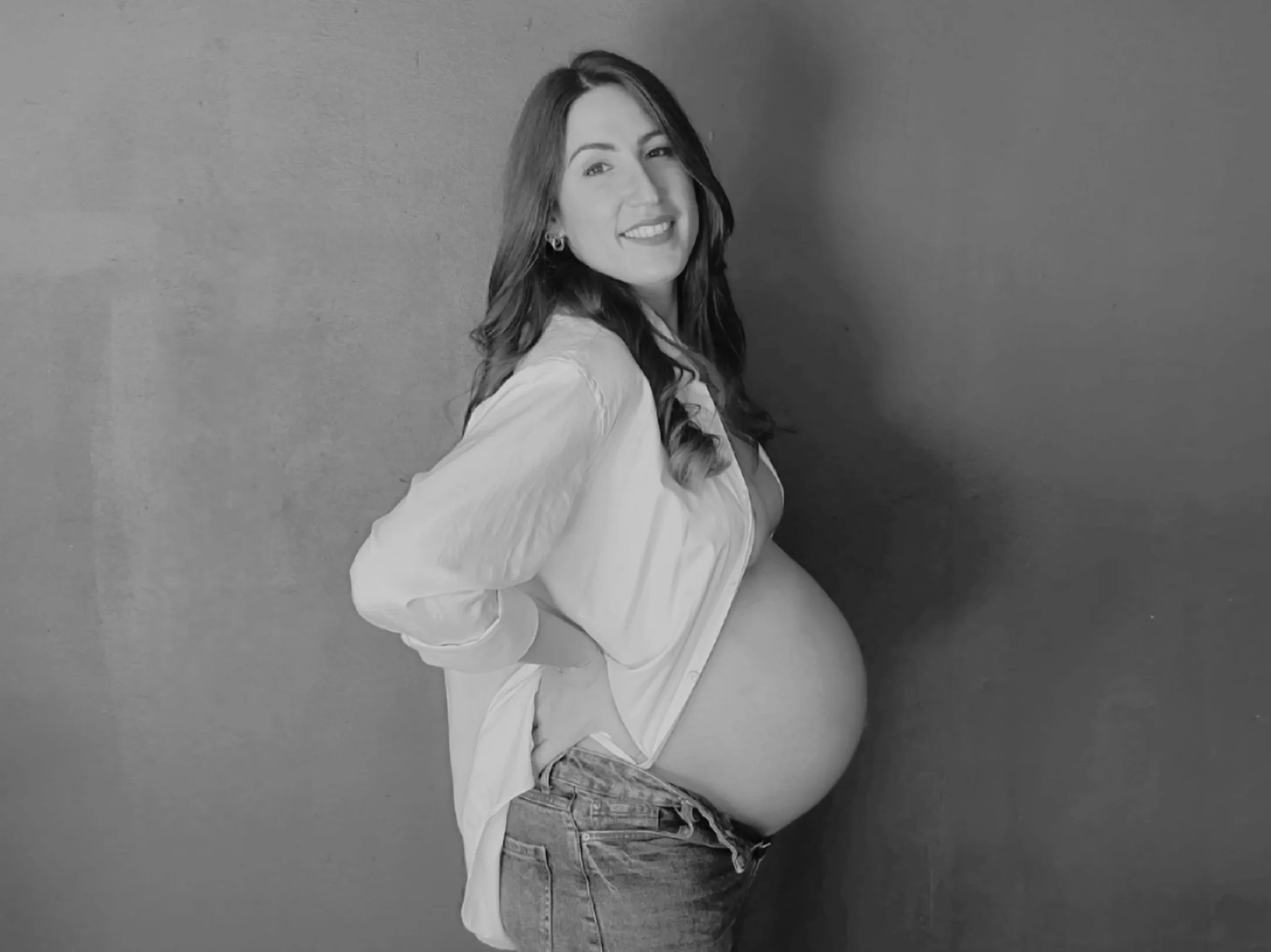Pregnant woman with long hair, a white shirt, jeans. and hands on her hips.