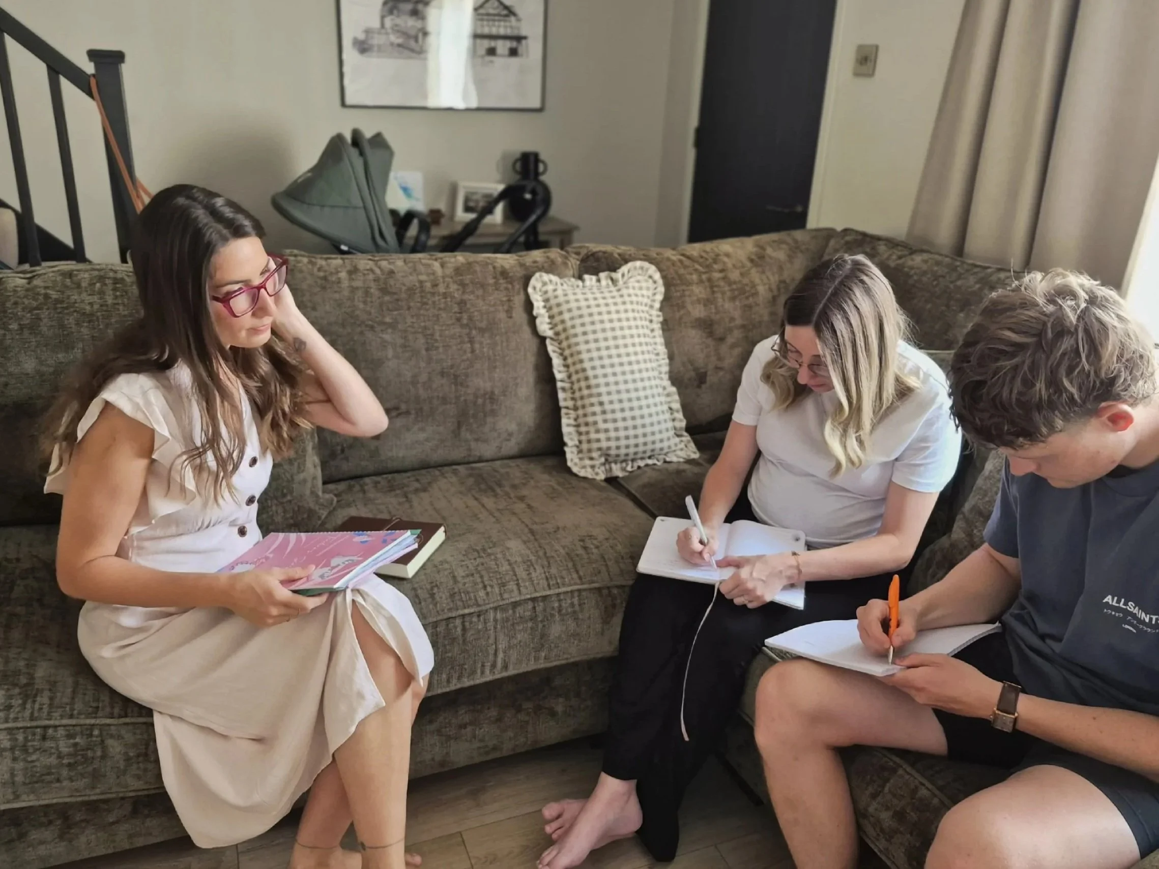 Four people sitting on a couch with notebooks and papers, some writing, in a living room with curtain on the right and a framed artwork on the wall.