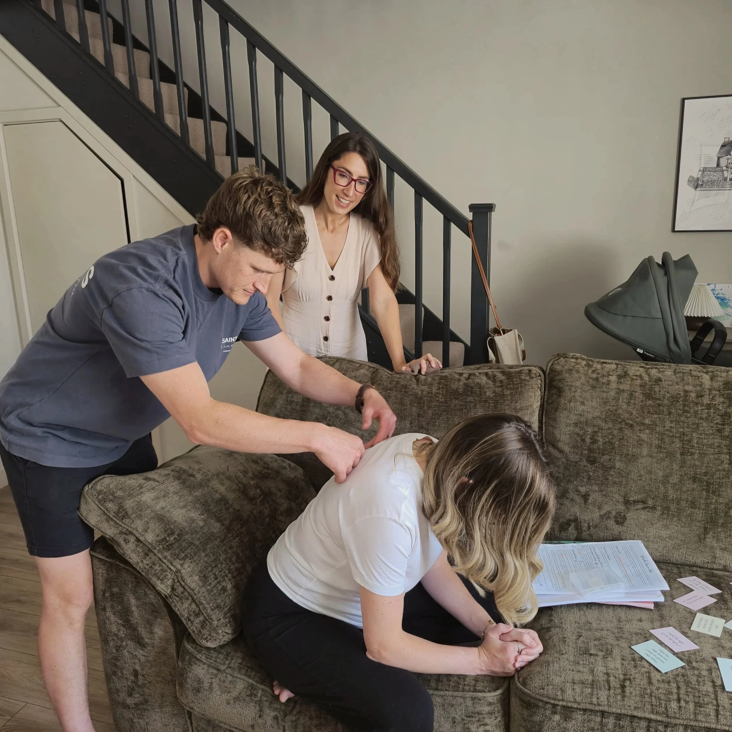 A pregnant woman is sitting on a couch, while her birth partner practises massage techniques on her back and the hypnobirthing teacher stands nearby giving guidance.