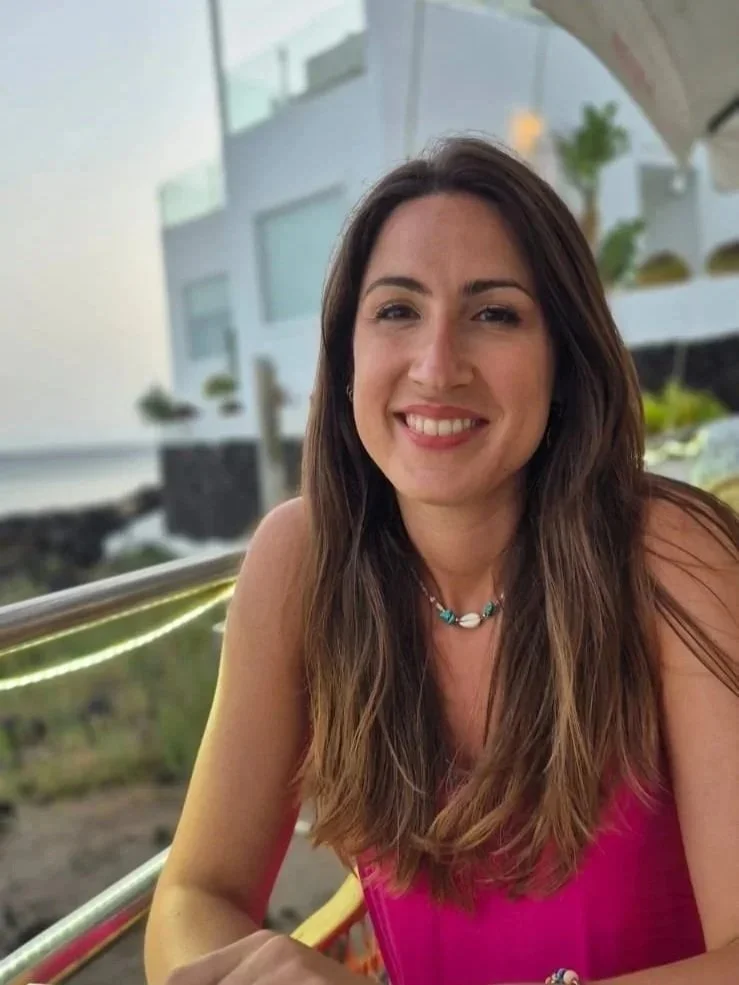 A woman with long brown hair smiling while sitting outdoors on a balcony, with buildings, greenery, and a coastal landscape in the background.
