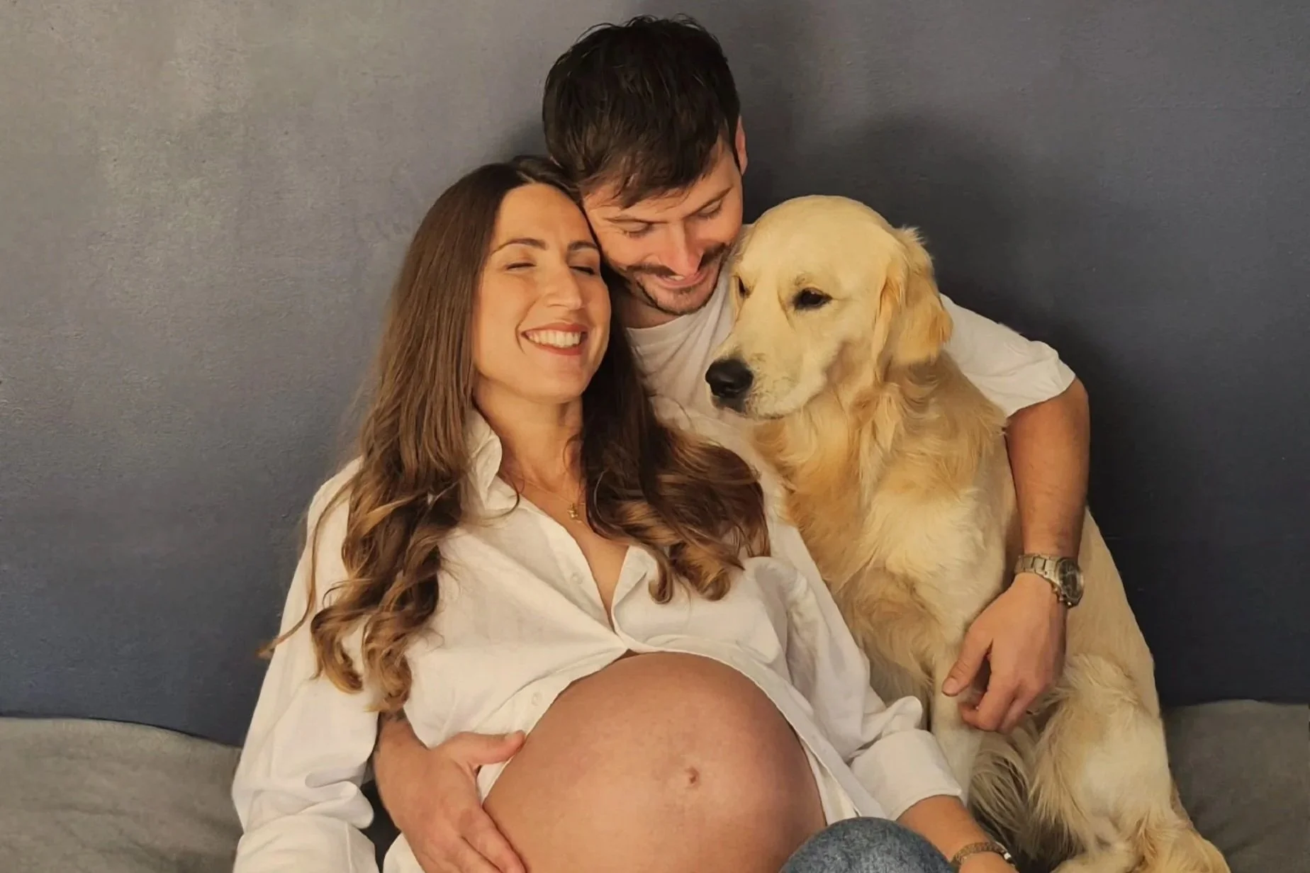 Pregnant person sitting alongside a man and a golden retriever at a pregnancy photoshoot.