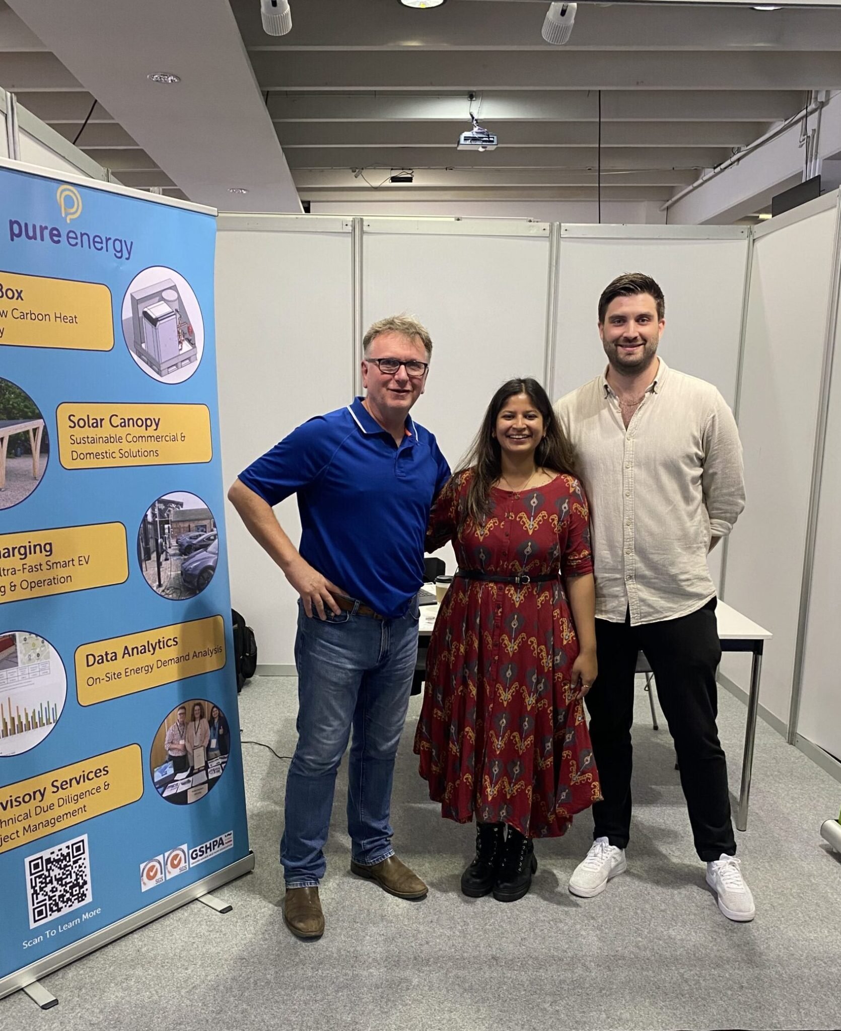 Three people standing together at an indoor event, smiling for the photo. Two men and one woman, with a display banner about energy solutions visible on the left side of the image.