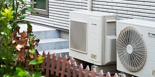 Outdoor air conditioning units beside a brick house and a small brown fence with greenery.