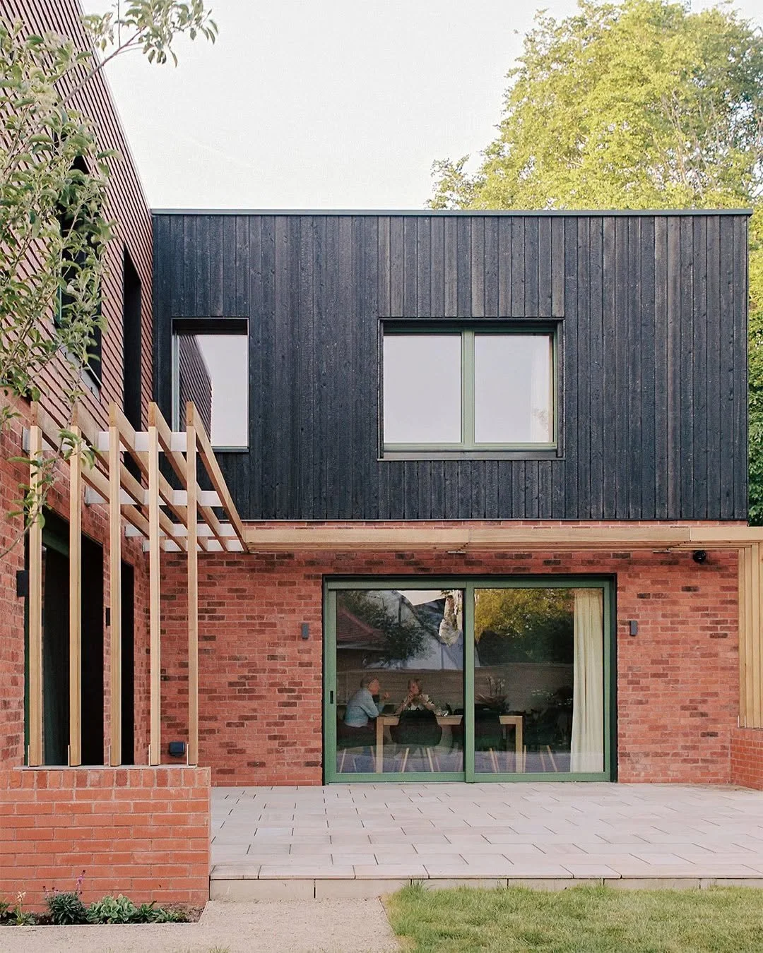 Modern residential house exterior with red brick on the lower level, black wooden on the upper level, and large glass sliding doors and windows, with a backyard patio and some trees in the background.