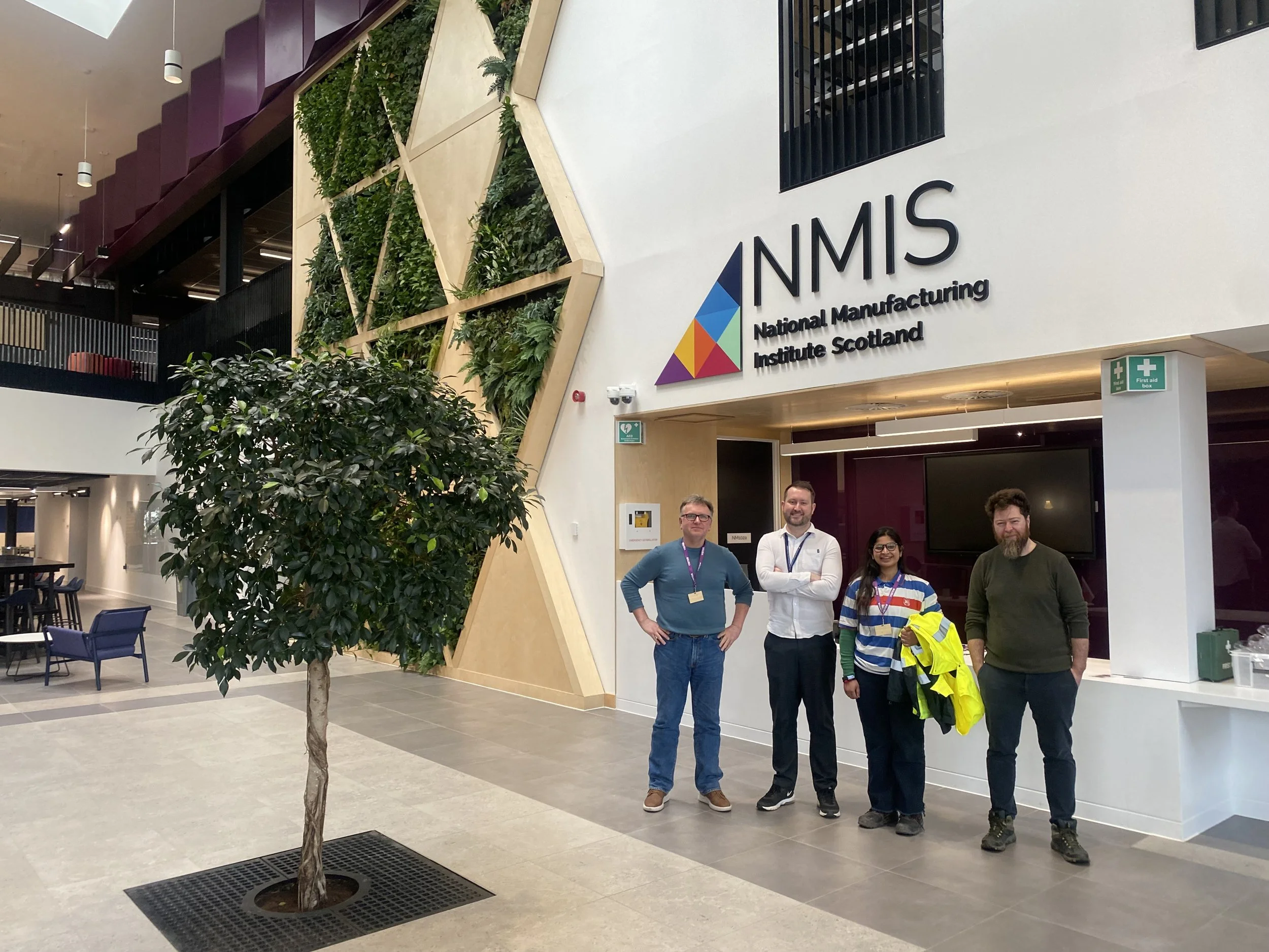 Four people are standing in front of a sign that reads 'NMS, National Manufacturing Institute Scotland' inside a modern building with green plants, seating area, and a staircase.