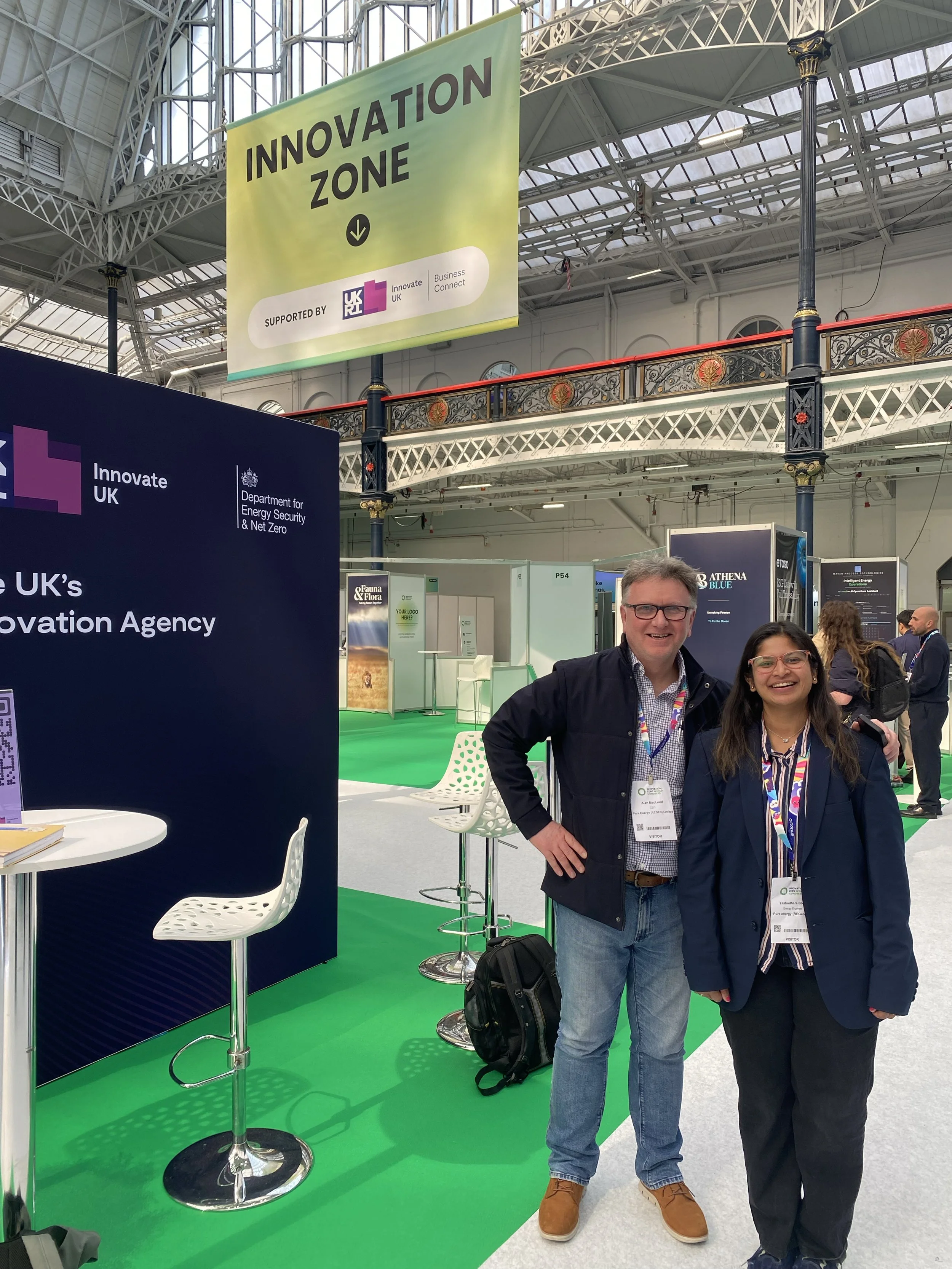 Two people smiling and standing at an exhibition booth with a sign that reads 'Innovation Zone'. The booth has white chairs and a standing table with books. A large banner overhead displays support from the UK government and Innovate UK.