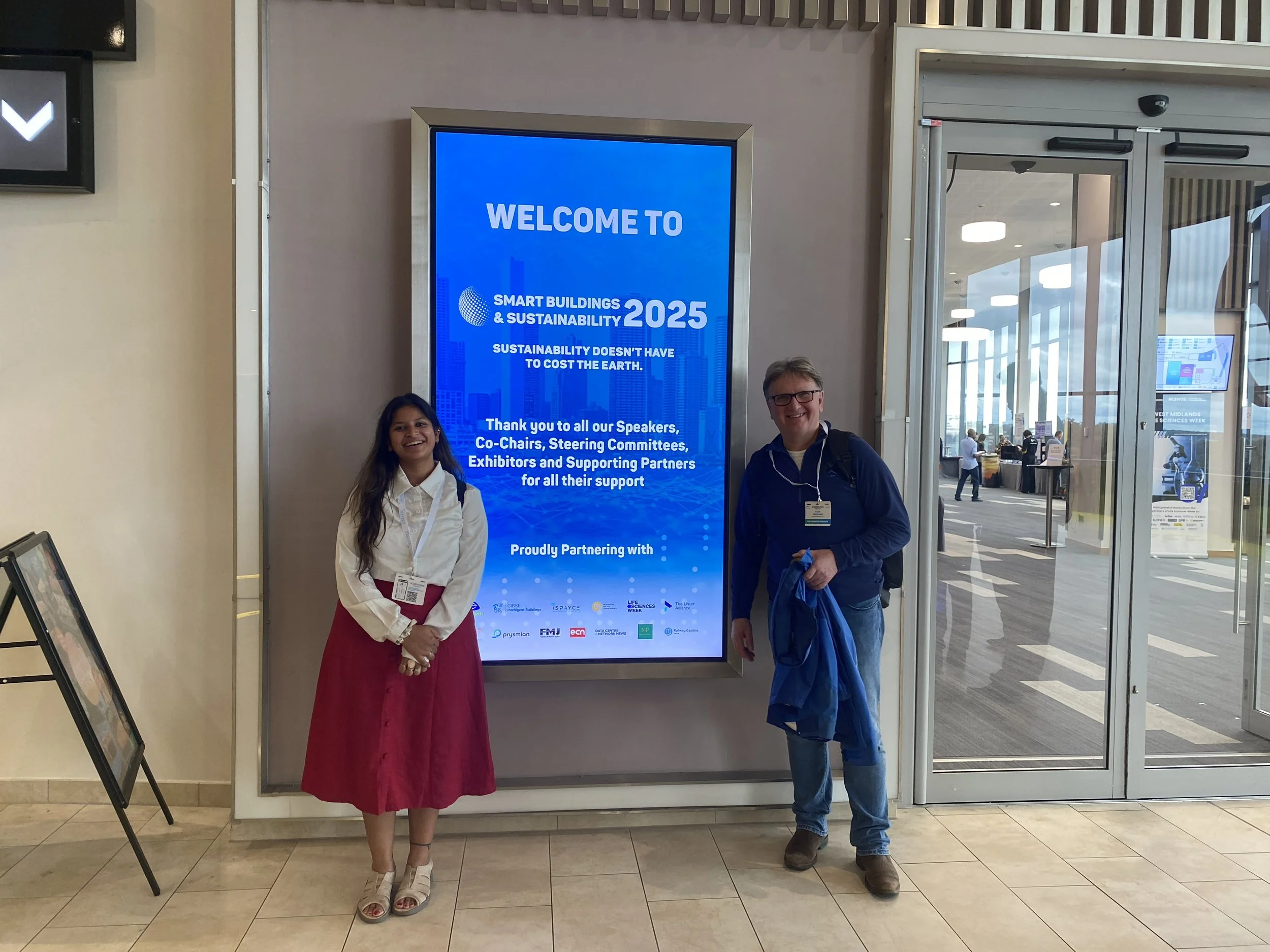 Two people standing in front of a large digital screen at a conference. The screen displays a welcome message for Smart Buildings & Sustainability 2025 event. The woman on the left is wearing a white blouse and a red skirt, and the man on the right is wearing glasses, a blue jacket, and a backpack. They are smiling and posing for the photo in a spacious, modern venue with glass doors and a bright interior.