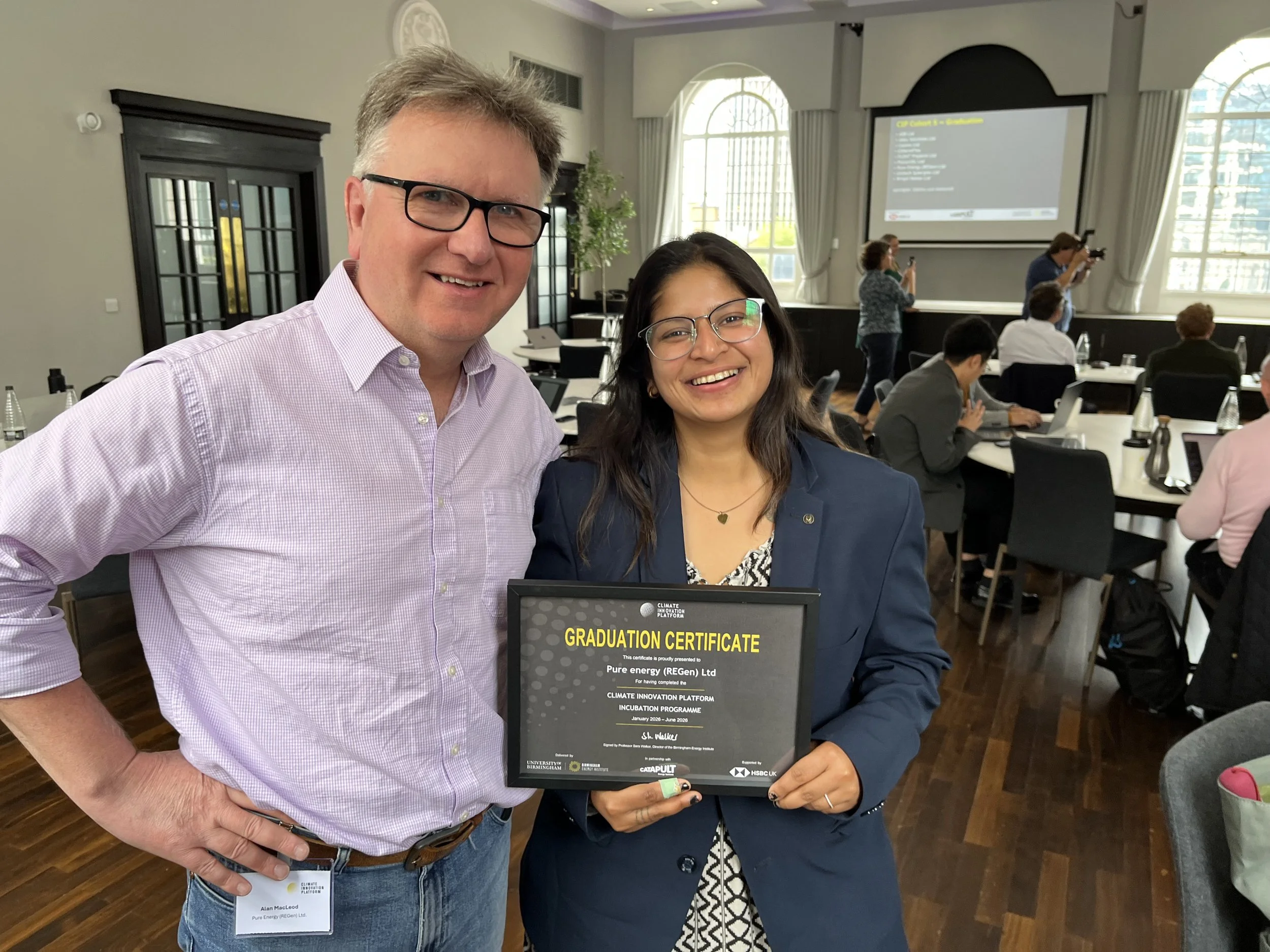A man and a woman smiling at the camera, holding a framed certificate that reads 'Graduation Certificate' for completing a climate innovation incubation program, in a conference room with attendees in the background.
