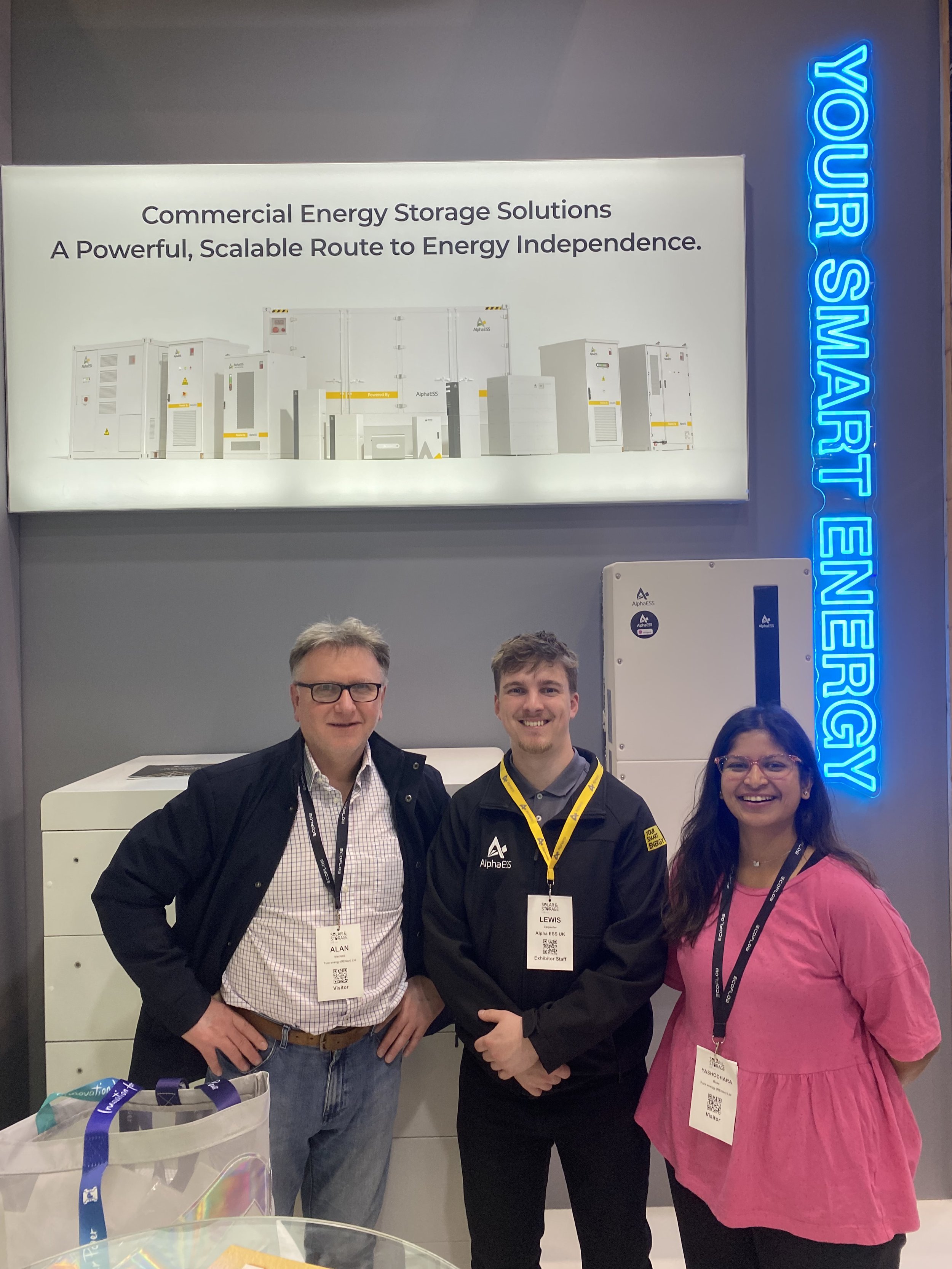 Three people, two men and one woman, standing indoors in front of an energy storage solutions display, smiling. The man on the left is wearing glasses and a checkered shirt, the man in the middle is wearing a black jacket with an AlphaESS logo, and the woman on the right is wearing a pink dress. They are at a conference or a trade show with name badges hanging around their necks.
