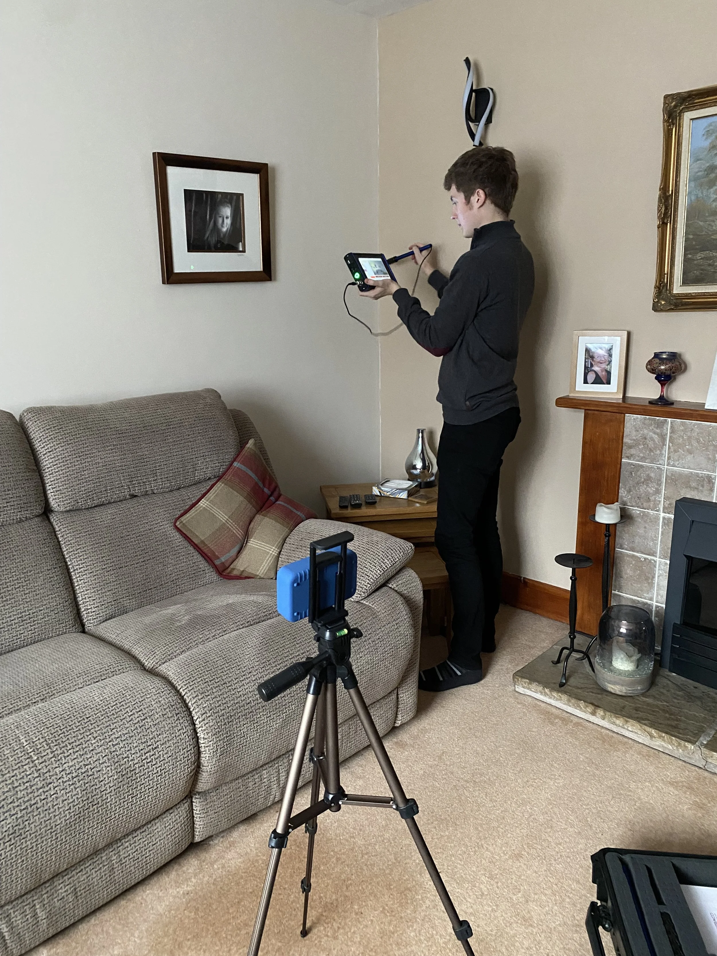A young man in a dark hoodie and black pants is standing in a living room, looking at a tablet device while holding a selfie stick. There is a tripod with a camera or phone set up in front of him, and a sofa with a cushion, a small wooden table, and a fireplace with a framed photo and decorative items on the mantel. The room is decorated with framed pictures on the walls, including a black-and-white portrait of a woman and a landscape painting.