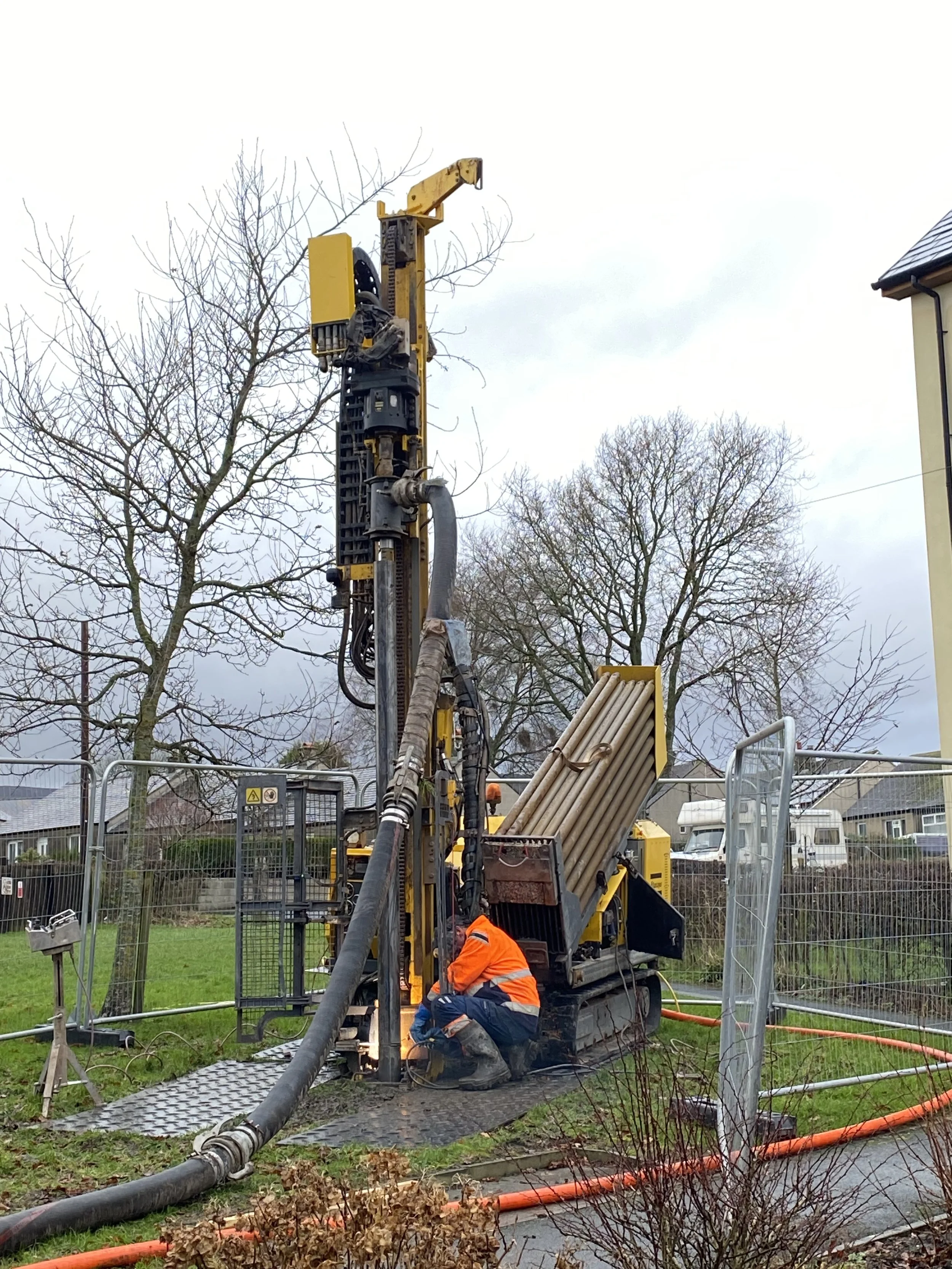 Worker in an orange safety jacket kneeling at a construction site, working on underground pipes with a large drilling machine and equipment, surrounded by leafless trees and a metal fence.
