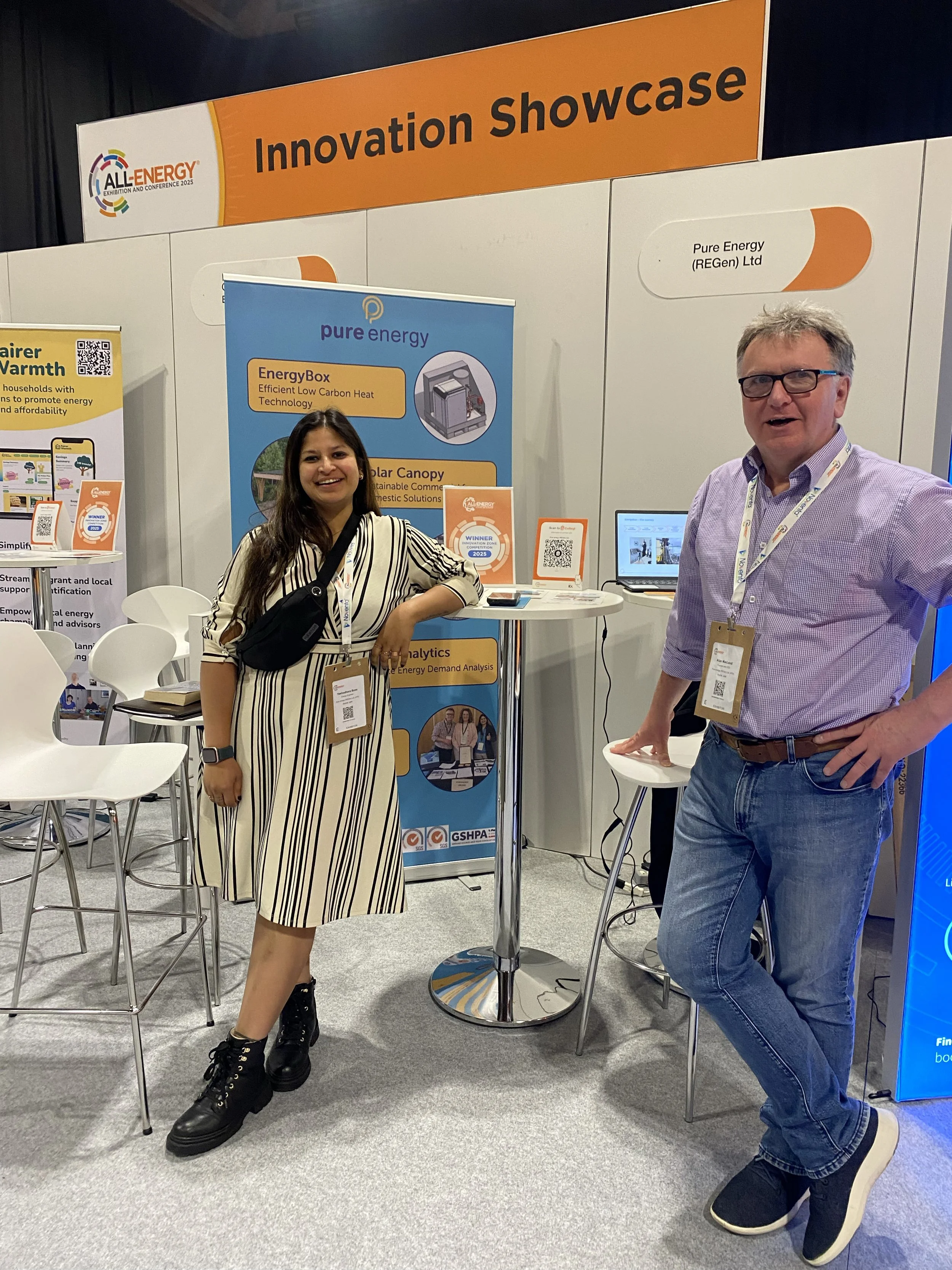 Two people standing at an exhibition booth labeled 'Innovation Showcase' at the All Energy conference 2025. A woman is smiling, wearing a black and white striped dress and black boots, standing next to a man in a light purple shirt and jeans, both with conference badges around their necks.