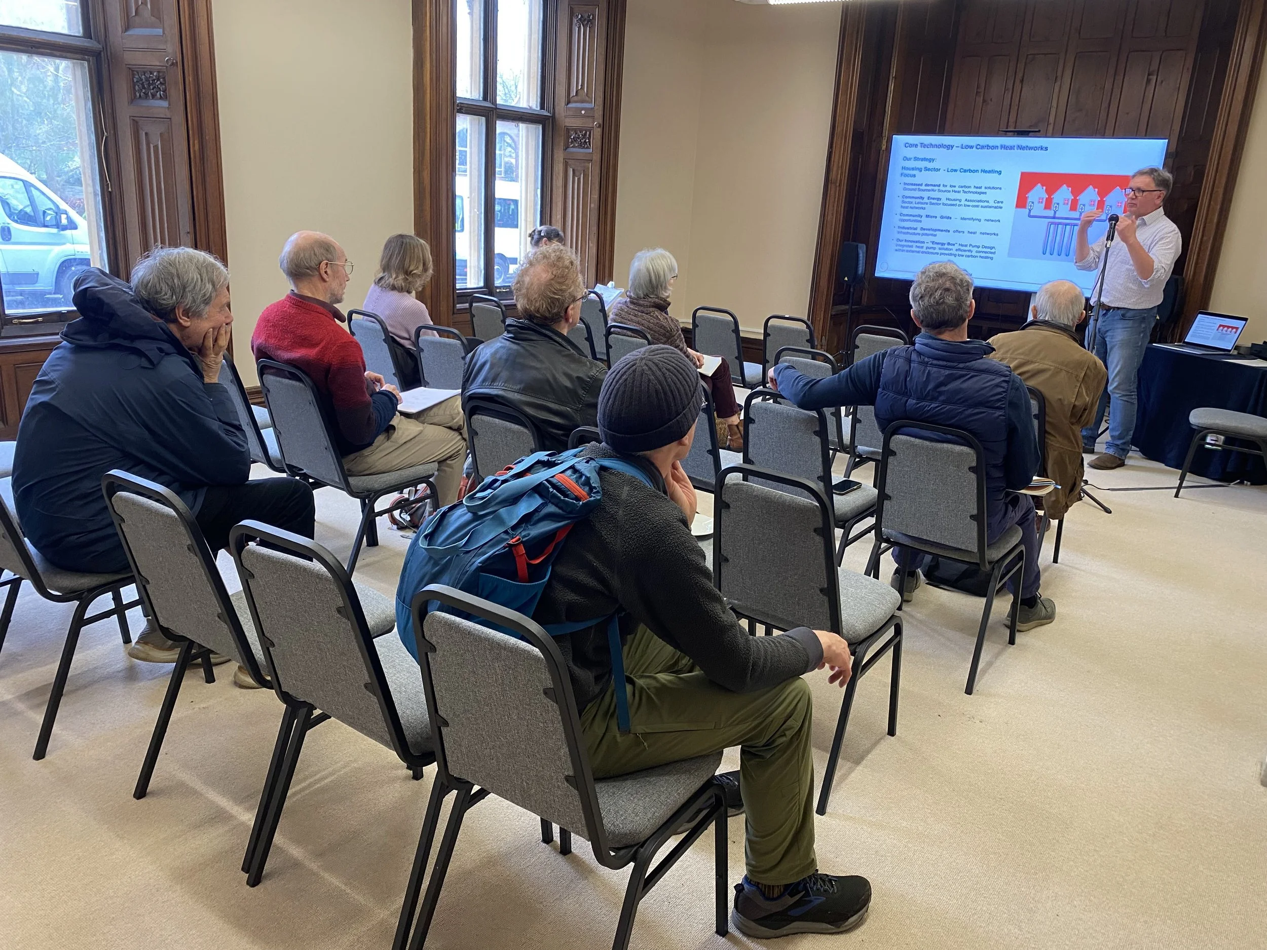 A man giving a presentation to an audience of adults seated in a room with large windows and wooden paneling. A large screen displays a slide titled 'Core Technology — Low Carbon Heat Networks.'