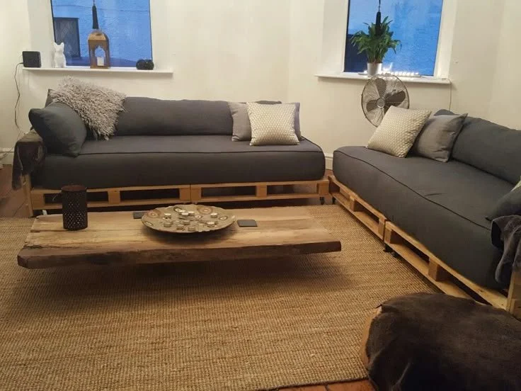 Living room with two gray couches made from wooden pallets, a wooden coffee table, a beige rug, and decorative pillows. There are windows with a potted plant and fan on the windowsill.