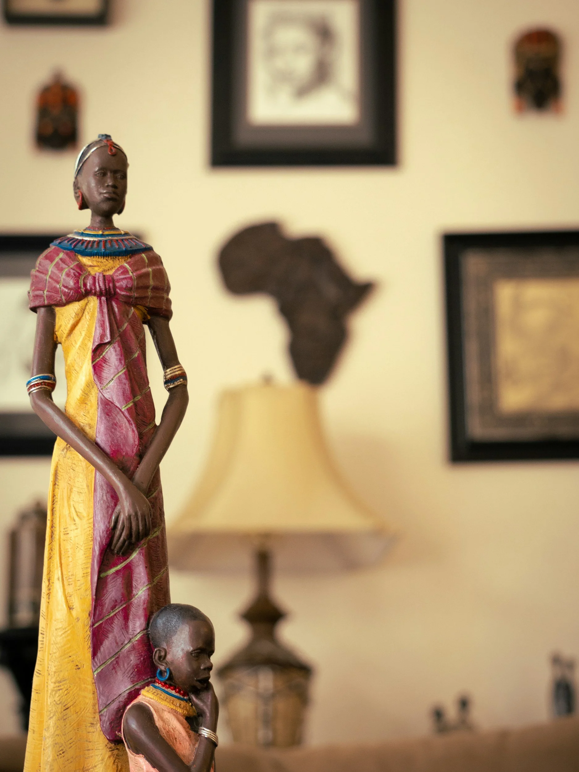 A close-up view of African figurines on display. One tall figurine in a pink and yellow traditional dress stands prominently, with another smaller figurine in a similar style just below it. The background features a beige wall with framed pictures and artifacts, some of which portray African maps and items.