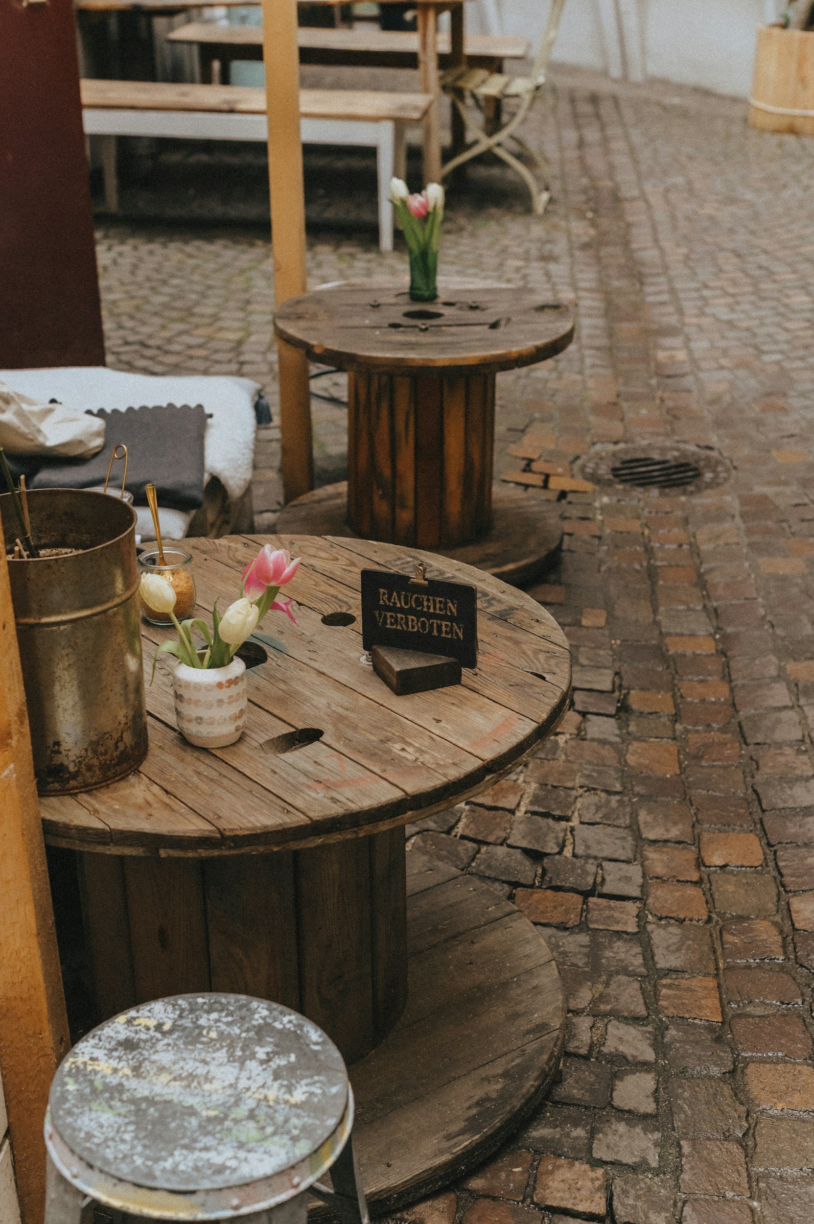 A small outdoor café or market stall with wooden barrels used as tables, decorated with flowers and small signs, on a cobblestone street.