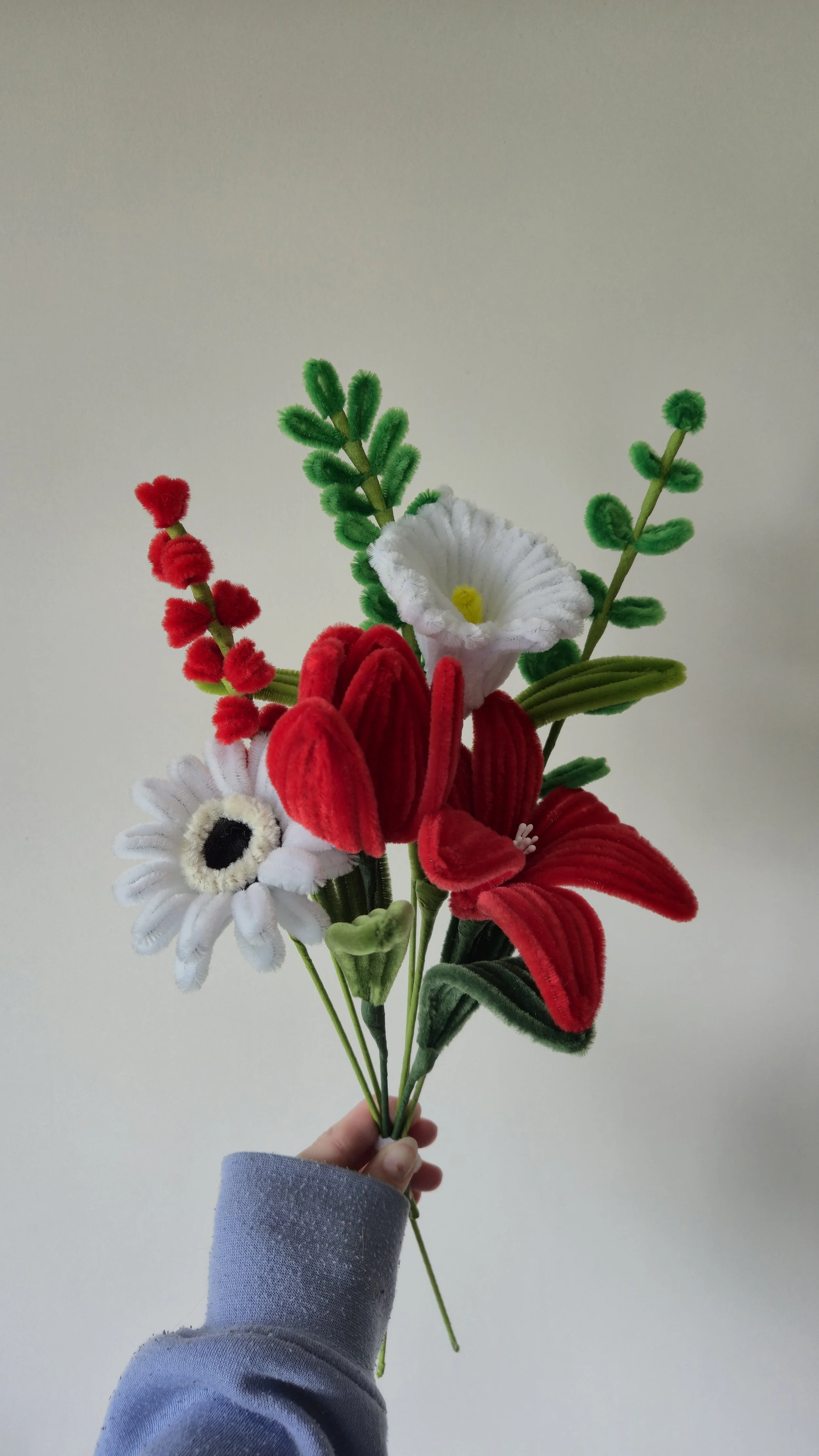 A hand holding a colorful artificial flower bouquet with red, white, and green elements against a plain white background.