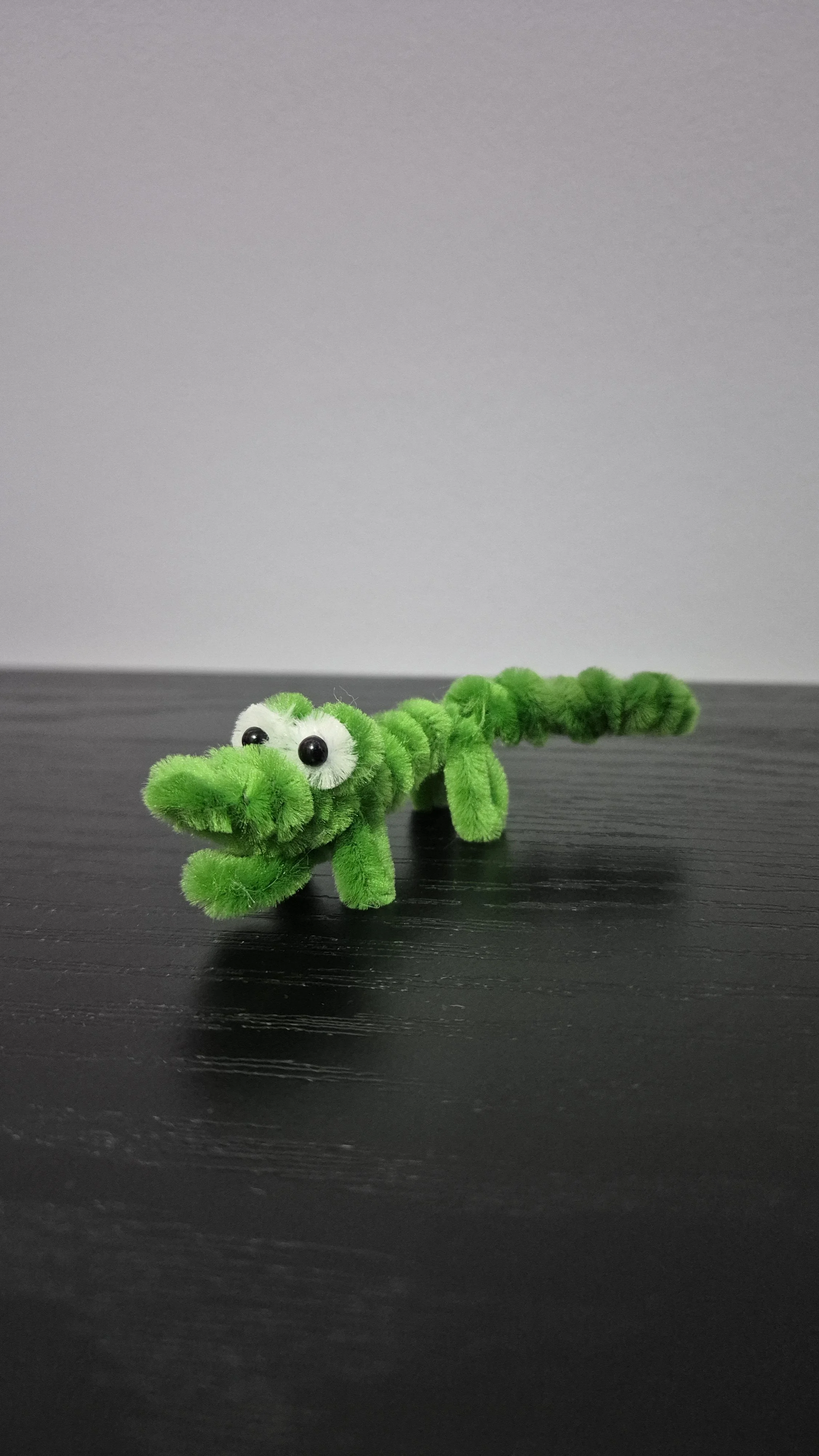 A small green plush toy shaped like a crocodile with googly eyes, sitting on a black surface against a gray background.