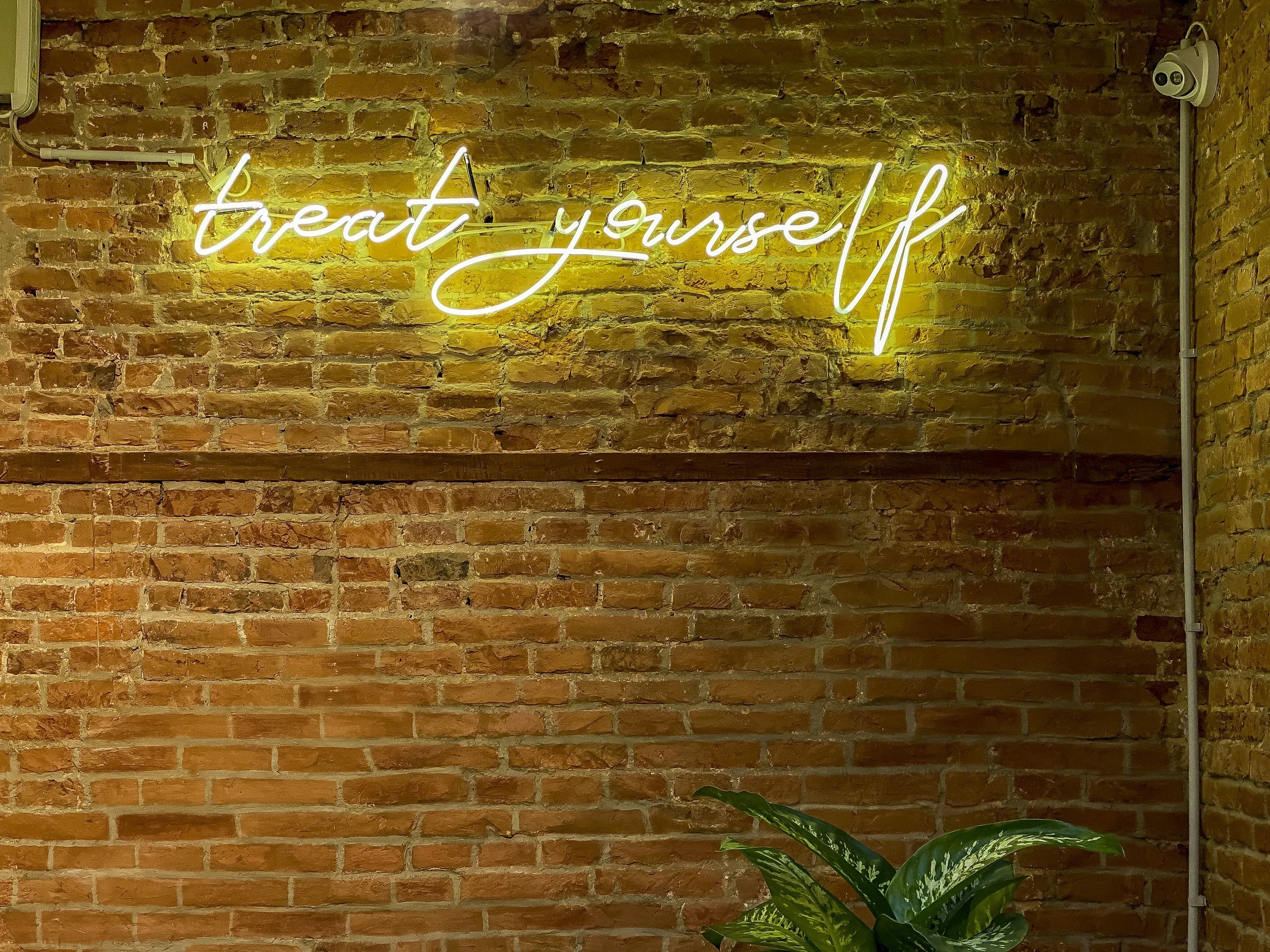 A neon sign that says 'treat yourself' in cursive white letters with yellow backlighting, mounted on a rustic brick wall, with some green plants at the bottom right corner.