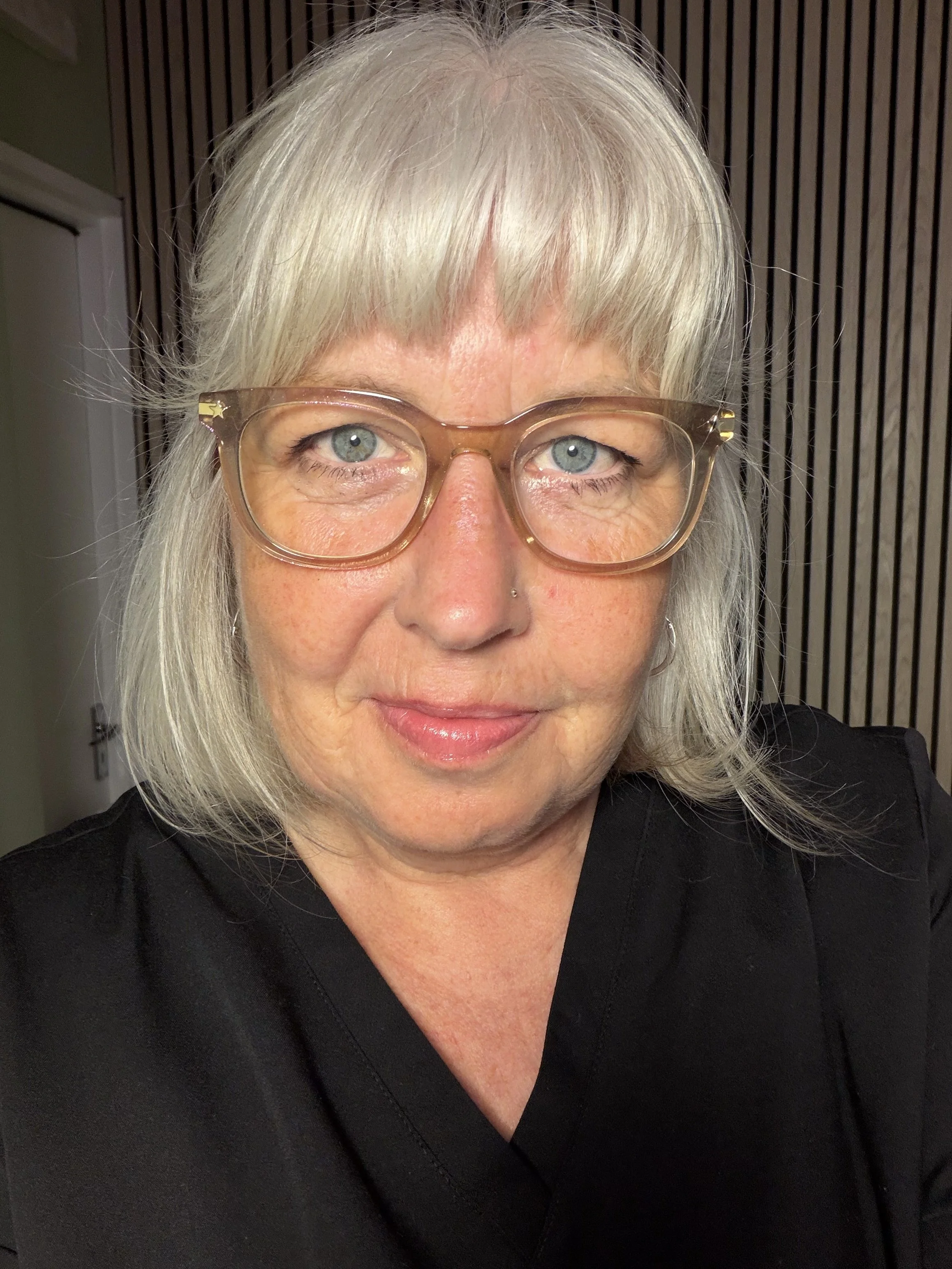 A  woman with long white hair, wearing large, light brown eyeglasses, smiling while wearing a black top, with a background of vertical wooden slats.