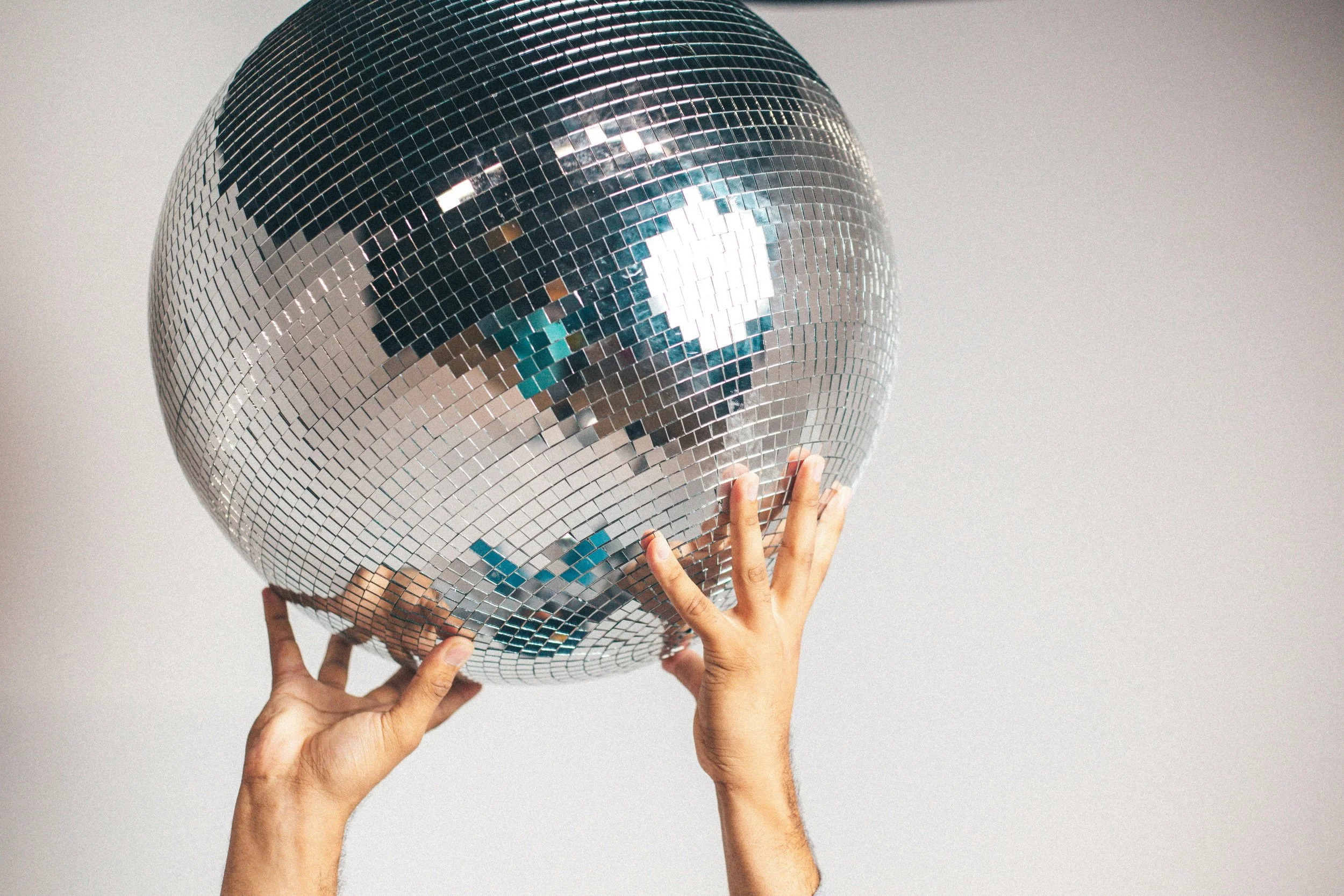 Multiple hands holding a disco ball decorated with small mirrored tiles.
