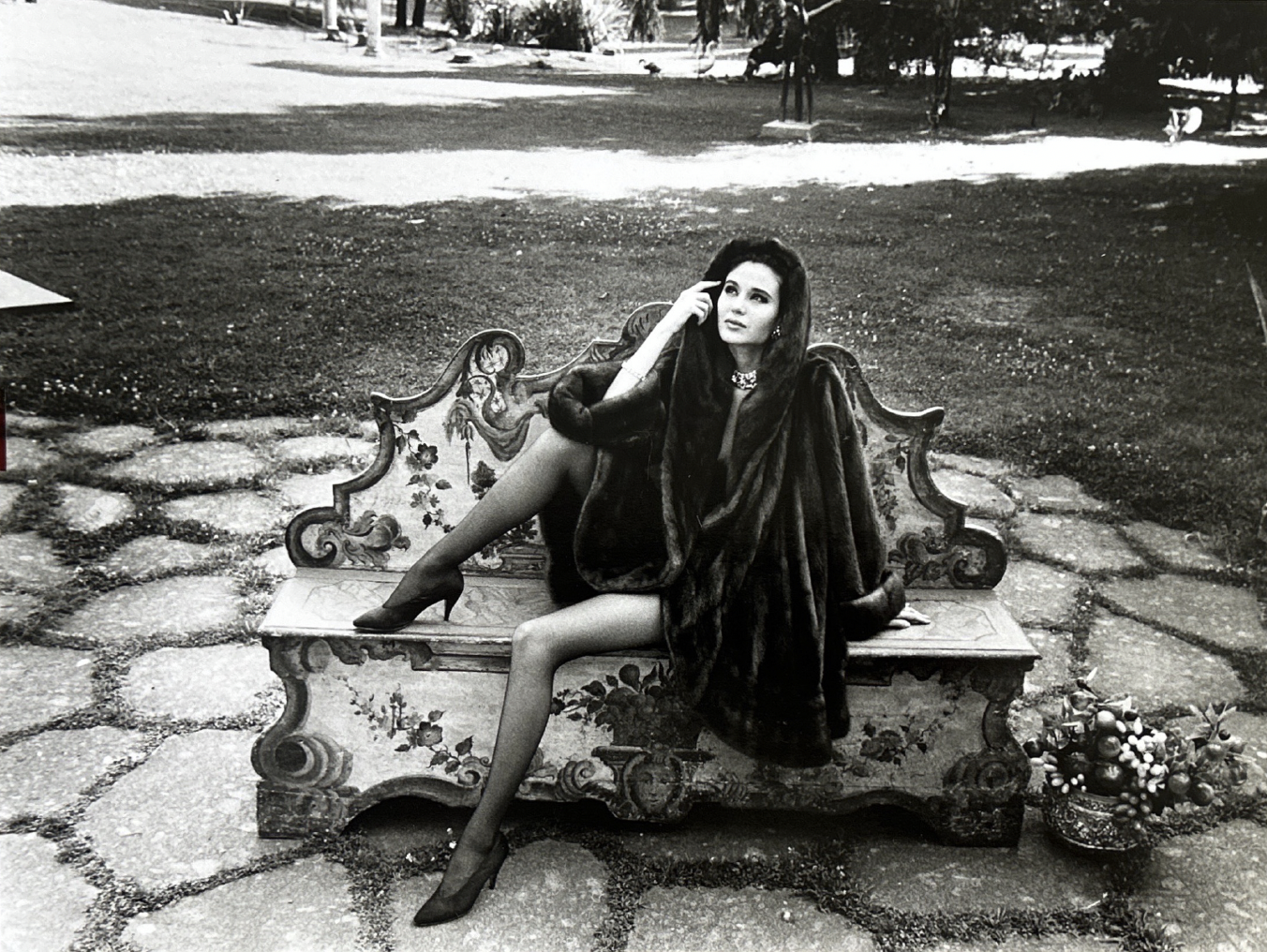 A woman sitting on an ornate bench outdoors, dressed in high heels and a fur coat, with a flower arrangement on the ground beside her.