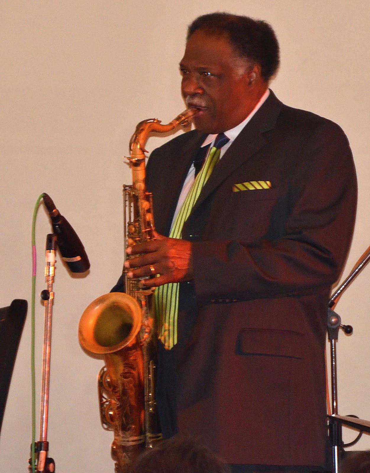 A man in a black suit and colorful striped tie playing a gold-colored saxophone on stage with a microphone nearby.