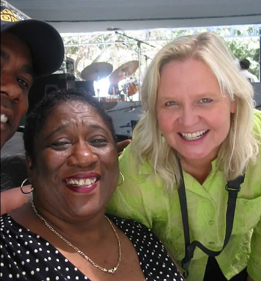 Three people smiling in a group photo during daytime, with a stage and musical instruments in the background.