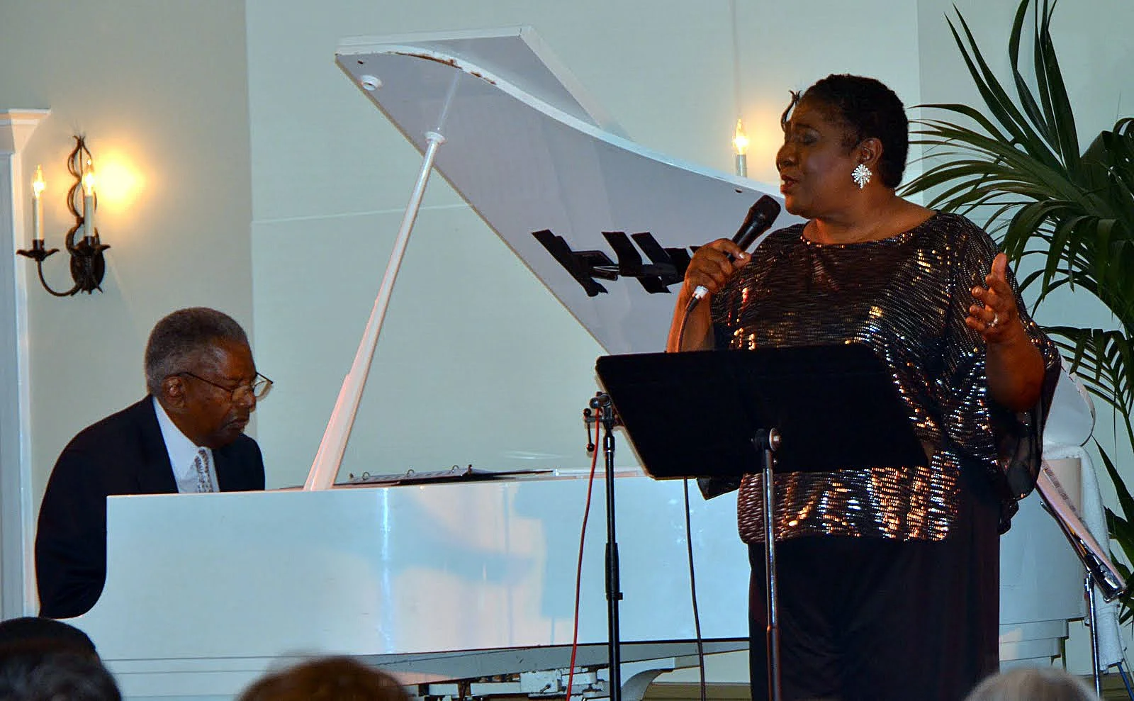 A woman in a sequined black top singing with a microphone, while a man in a suit plays the white grand piano beside her, in a decorated room with wall sconces and a large green plant.