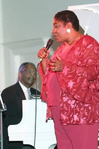 A woman in pink and red clothing speaking into a microphone, with a pianist in the background.