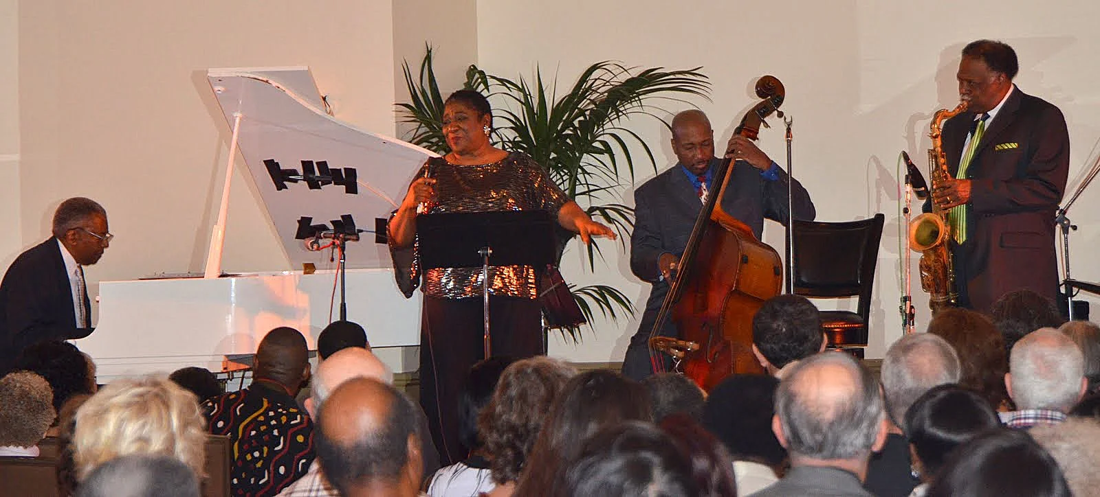 Jazz band performing live on stage with pianist, female vocalist, upright bassist, and saxophonist in front of seated audience.