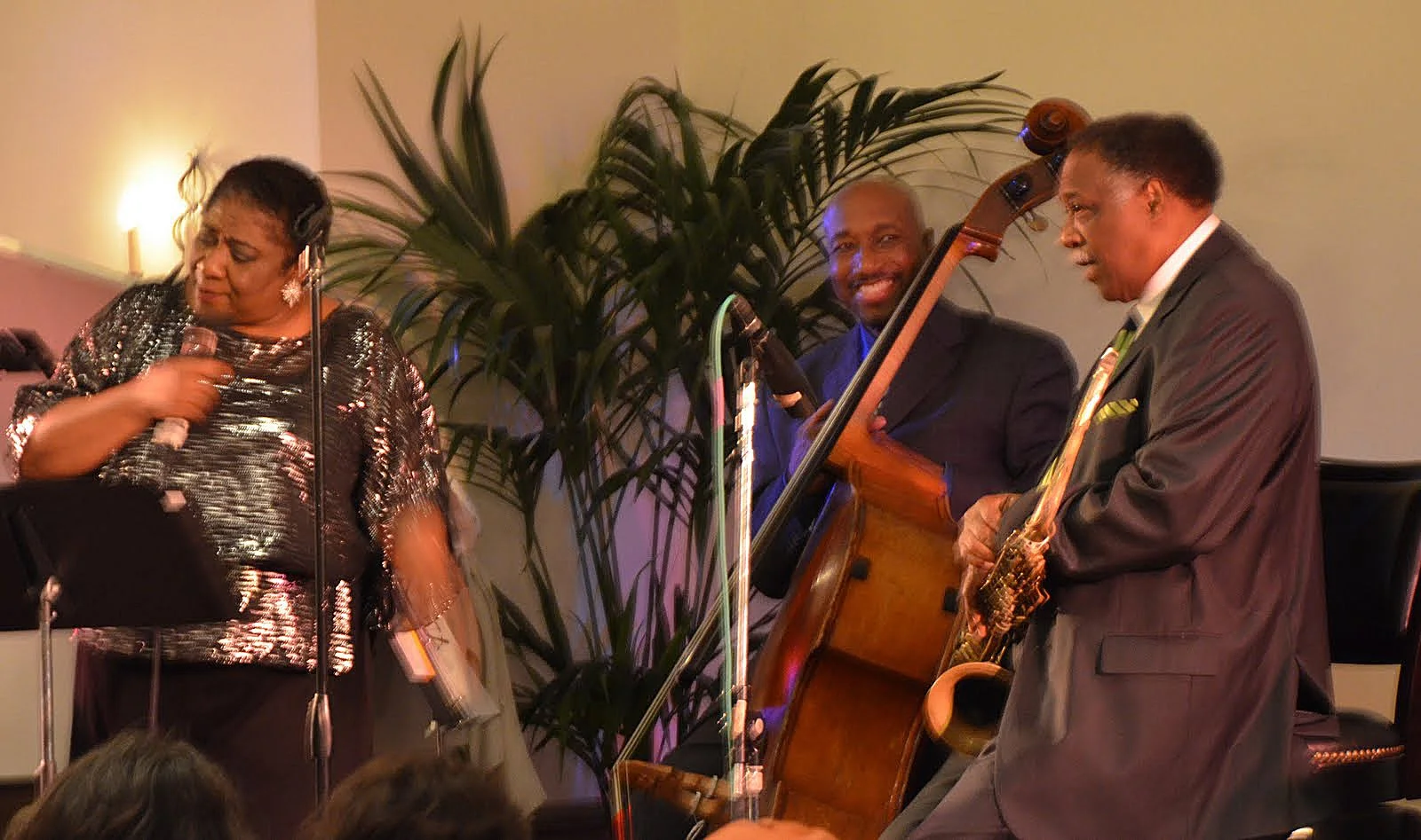 Jazz musicians performing on stage with a woman singing, a man playing double bass, and a man playing saxophone, with a large plant and dim lighting in the background.