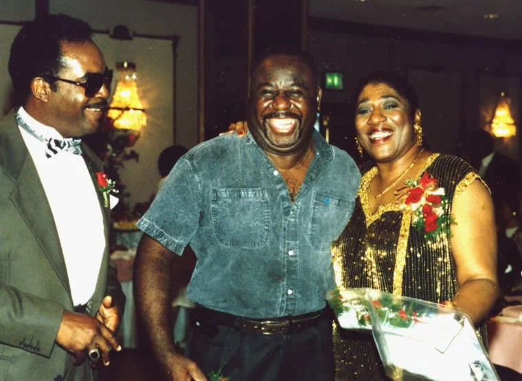 Three people smiling at a party, two men and one woman, with the man in the middle laughing, the woman holding a gift and flowers, decorations and dim lighting in the background.
