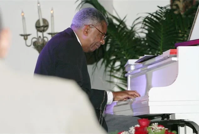 Man playing white piano with a serious expression, surrounded by plants and candles.