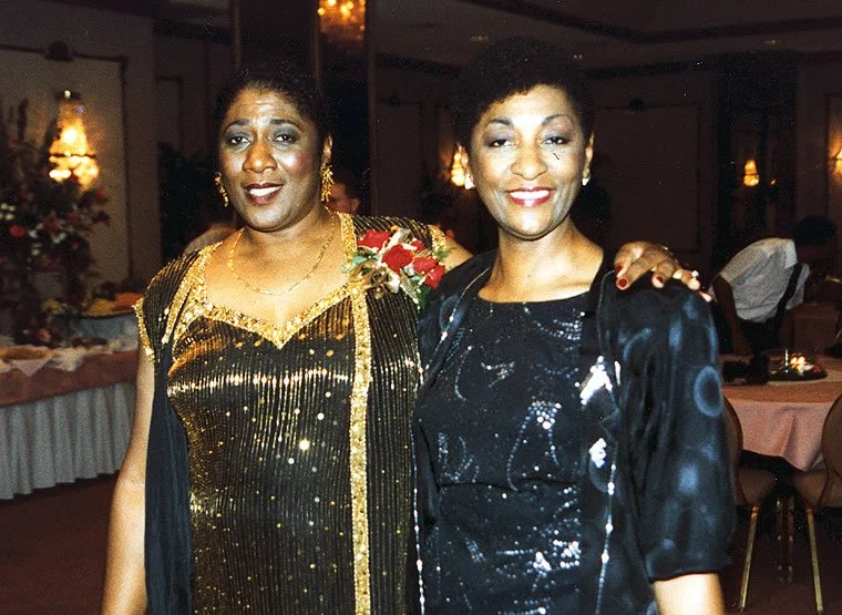 Two women in formal dresses posing together at an indoor event with decorated tables and warm lighting.