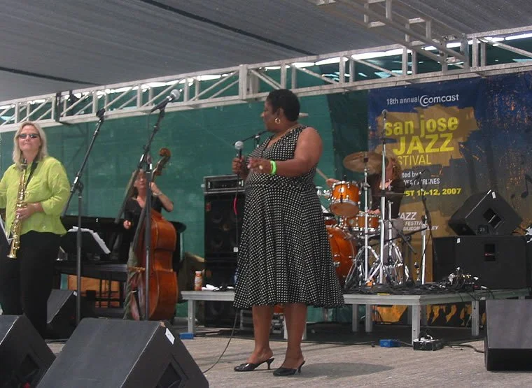 Female vocalist singing on stage at the San Jose Jazz Festival, with musicians playing instruments behind her, including a double bass and drums, during daytime.