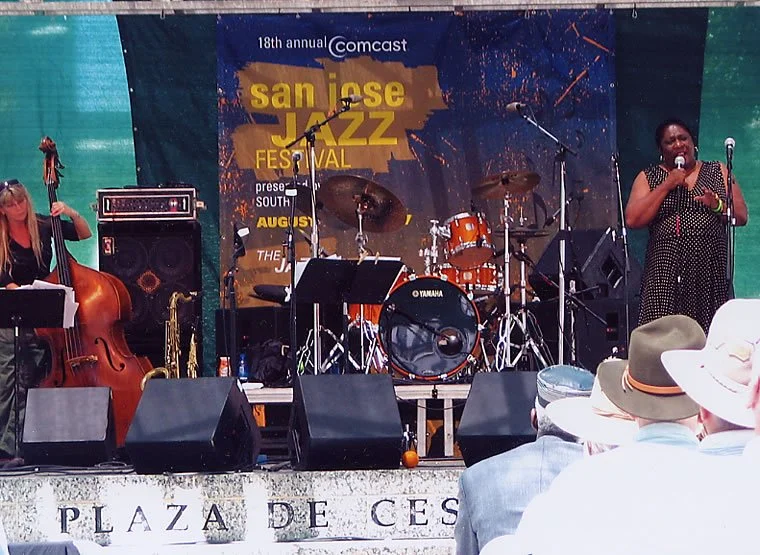 Musicians performing on stage at the San Jose Jazz Festival, with a large banner in the background and an audience watching, during daytime.