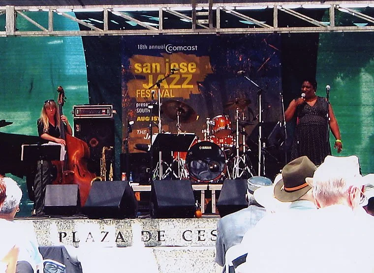 Musicians performing on stage at the San Jose Jazz Festival, with drum set, keyboard, double bass, and singer, during daytime outdoor event.