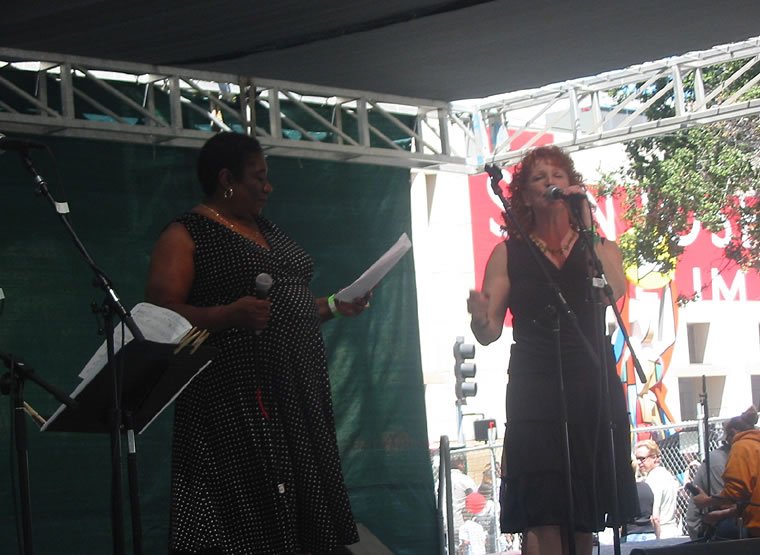 Two women speaking or singing on an outdoor stage, one holding a microphone and papers, the other speaking into a microphone, with musical equipment and a crowd in the background.